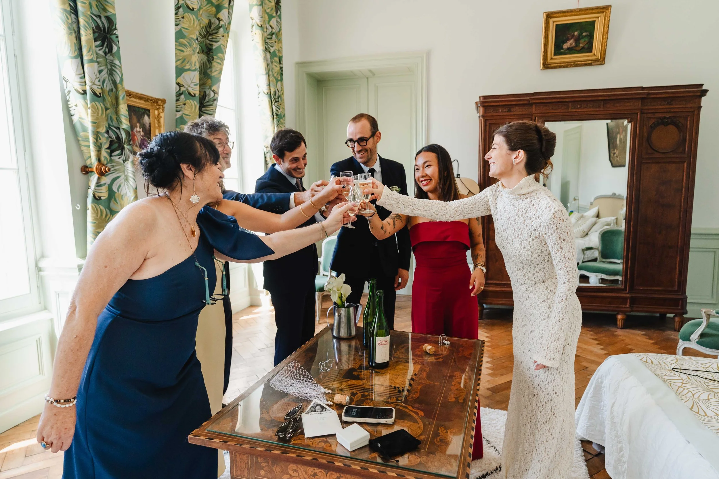 Groupe de personnes habillées élégamment pour un mariage, levant des coupes de champagne pour un toast dans une pièce décorée avec des meubles en bois et des draperies florales.
