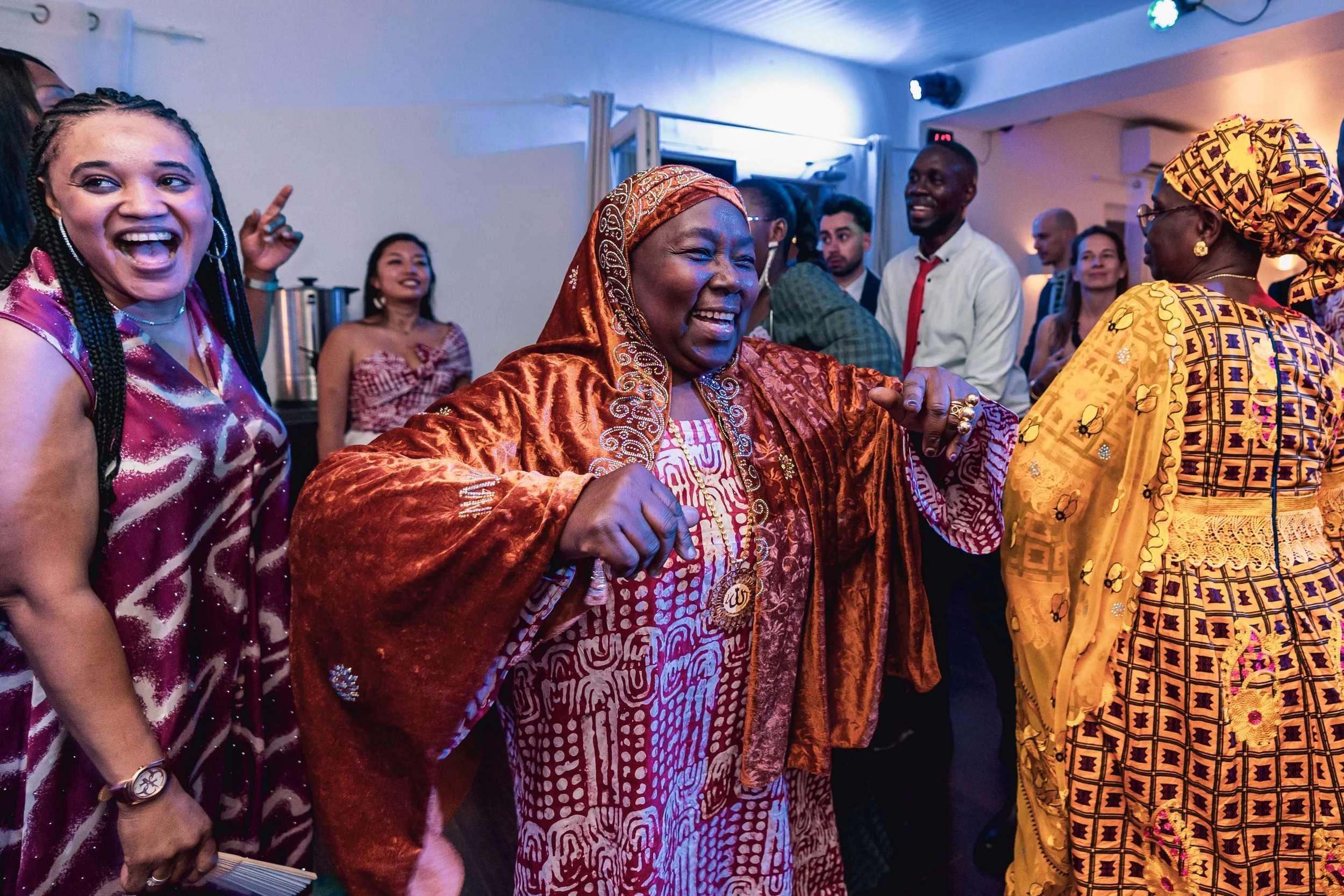 Groupe de personnes dansant et souriant lors d'une fête, vêtues de tenues traditionnelles africaines, dans une salle avec éclairage bleu et blanc.