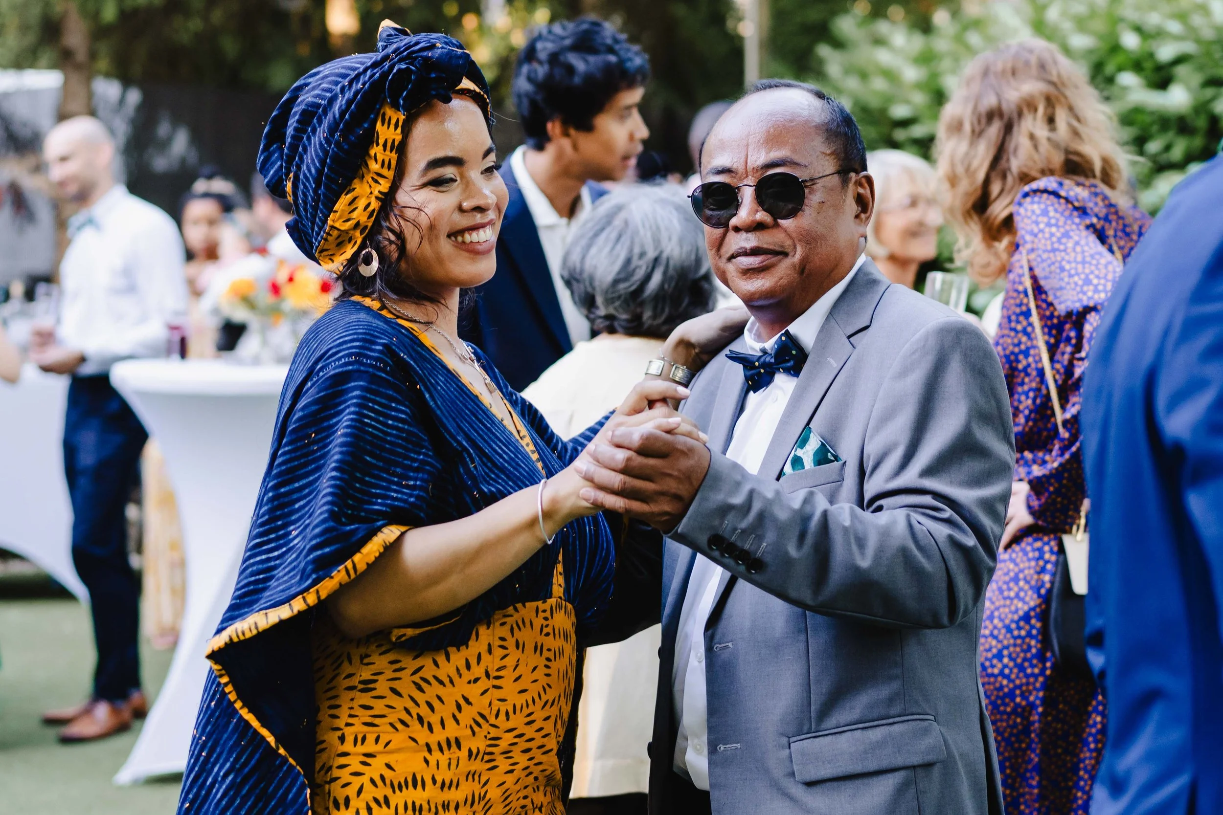 deux personnes dansant lors d'une fête en plein air, une femme portant une tenue traditionnelle colorée avec un turban et un homme en costume avec des lunettes noires