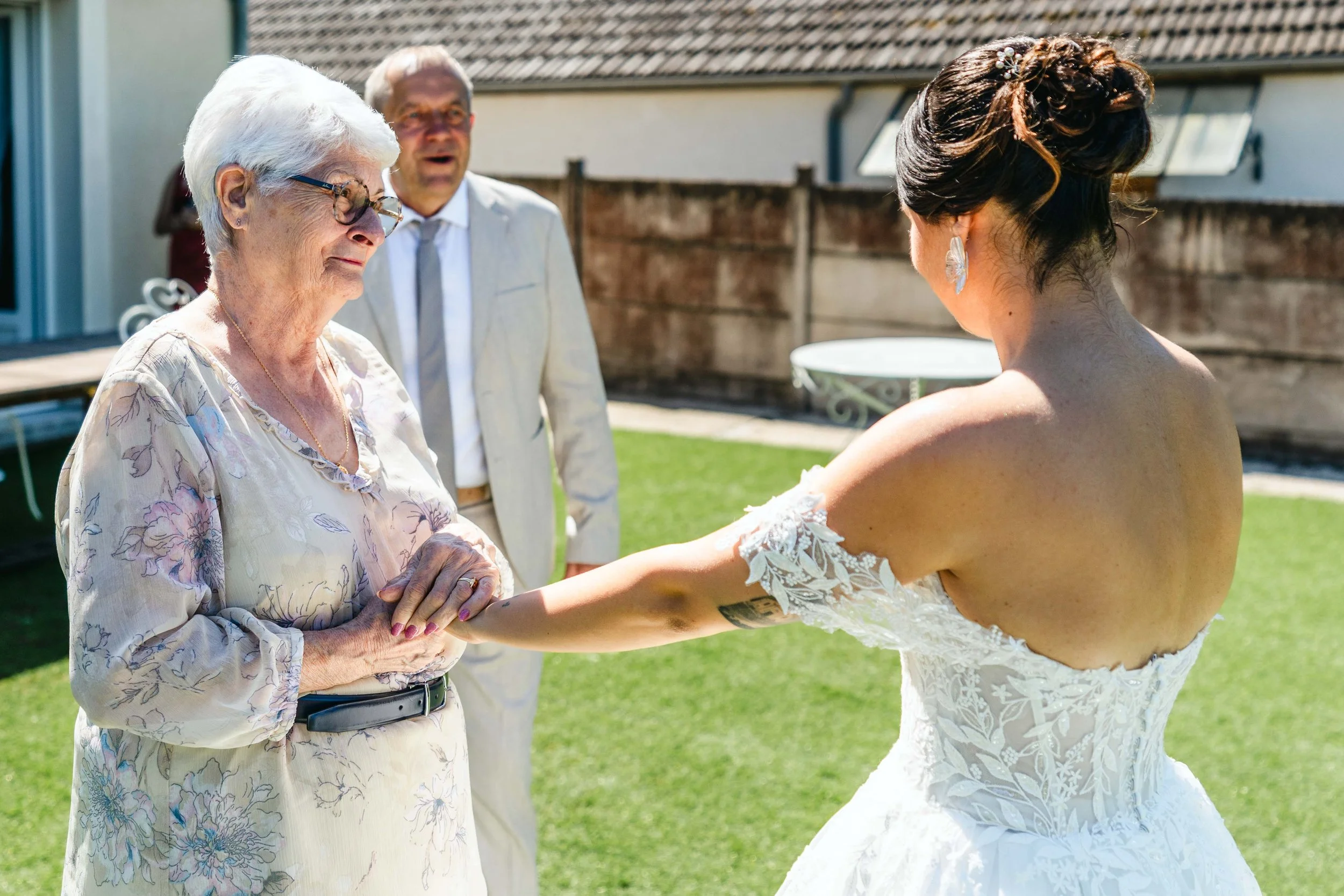 Une mariée en robe de mariage blanche tenant la main d'une femme âgée lors d'une cérémonie de mariage en plein air, avec un homme en veste blanche derrière eux.