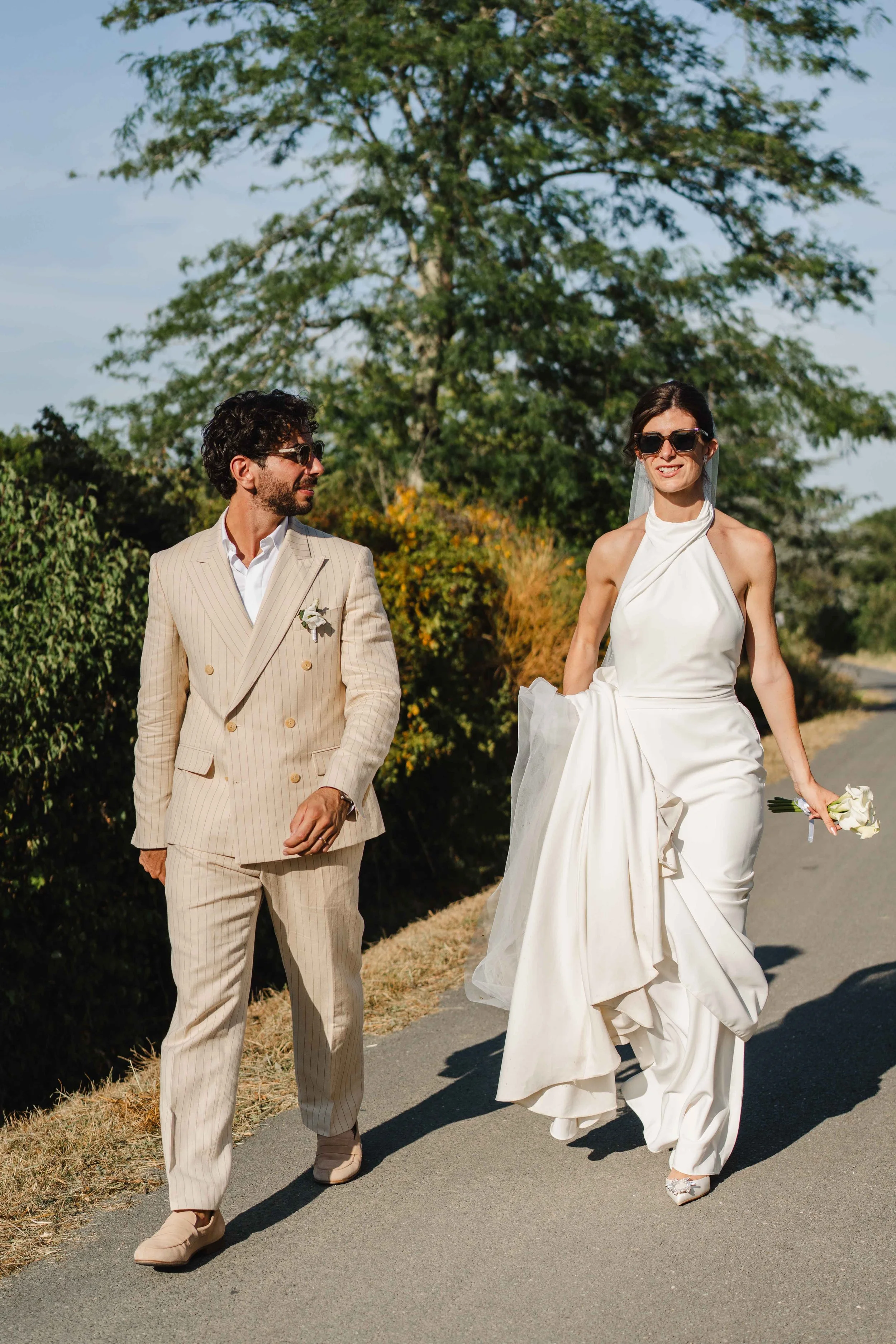 Un couple habillé en tenue de mariage marchant sur un chemin en plein air, au soleil, avec des arbres en arrière-plan.