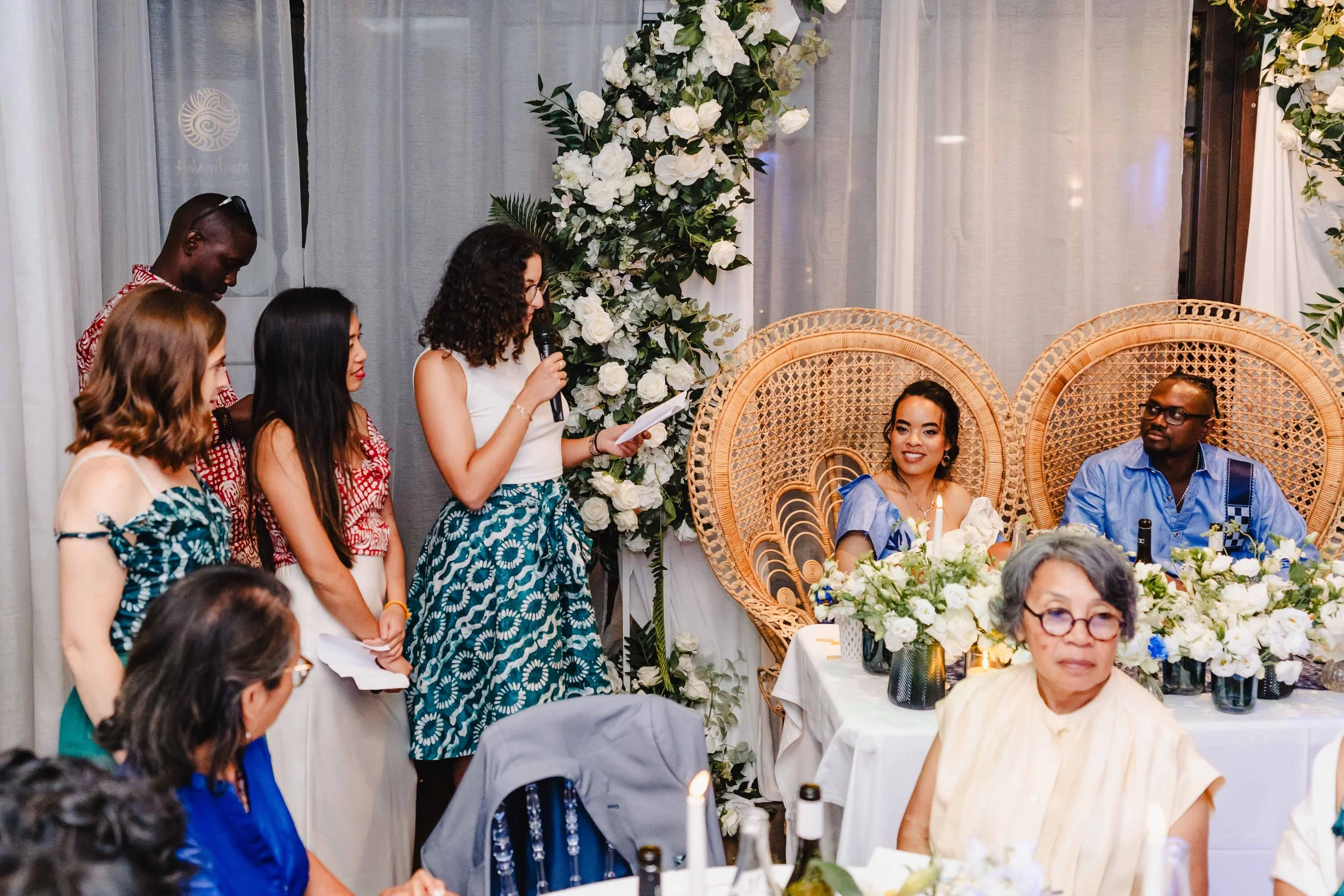Un groupe de personnes lors d'une cérémonie ou d'un discours, avec deux personnes assises dans des peignes en osier devant une décoration florale abondante.