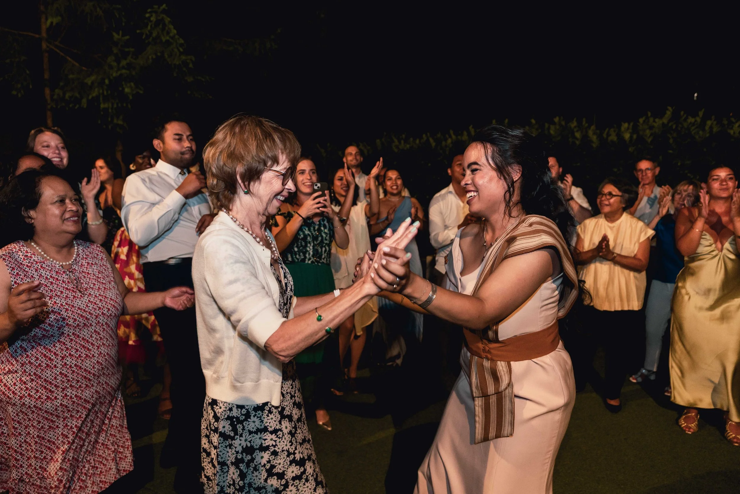 Deux femmes dansent et sourient lors d'une fête en plein air, entourées de personnes qui applaudissent et prennent des photos, la nuit.