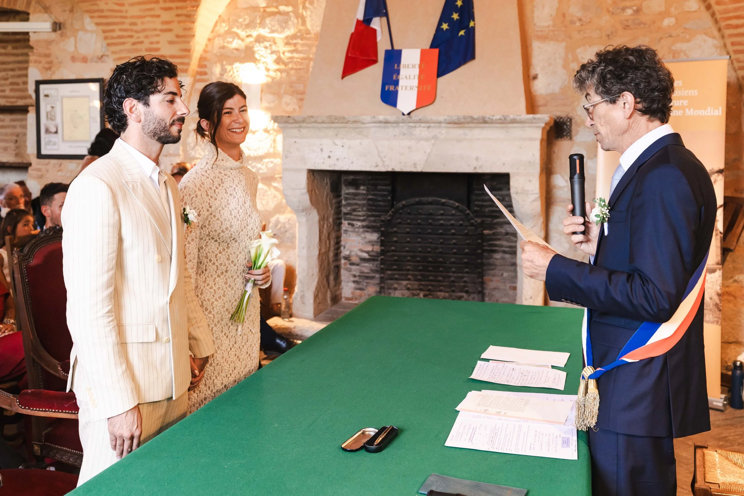Un couple marié lors de leur cérémonie de mariage civil en présence d'un officier d'État civil dans une salle avec des murs en pierre et un drapeau français et européen suspendus au-dessus de la cheminée.