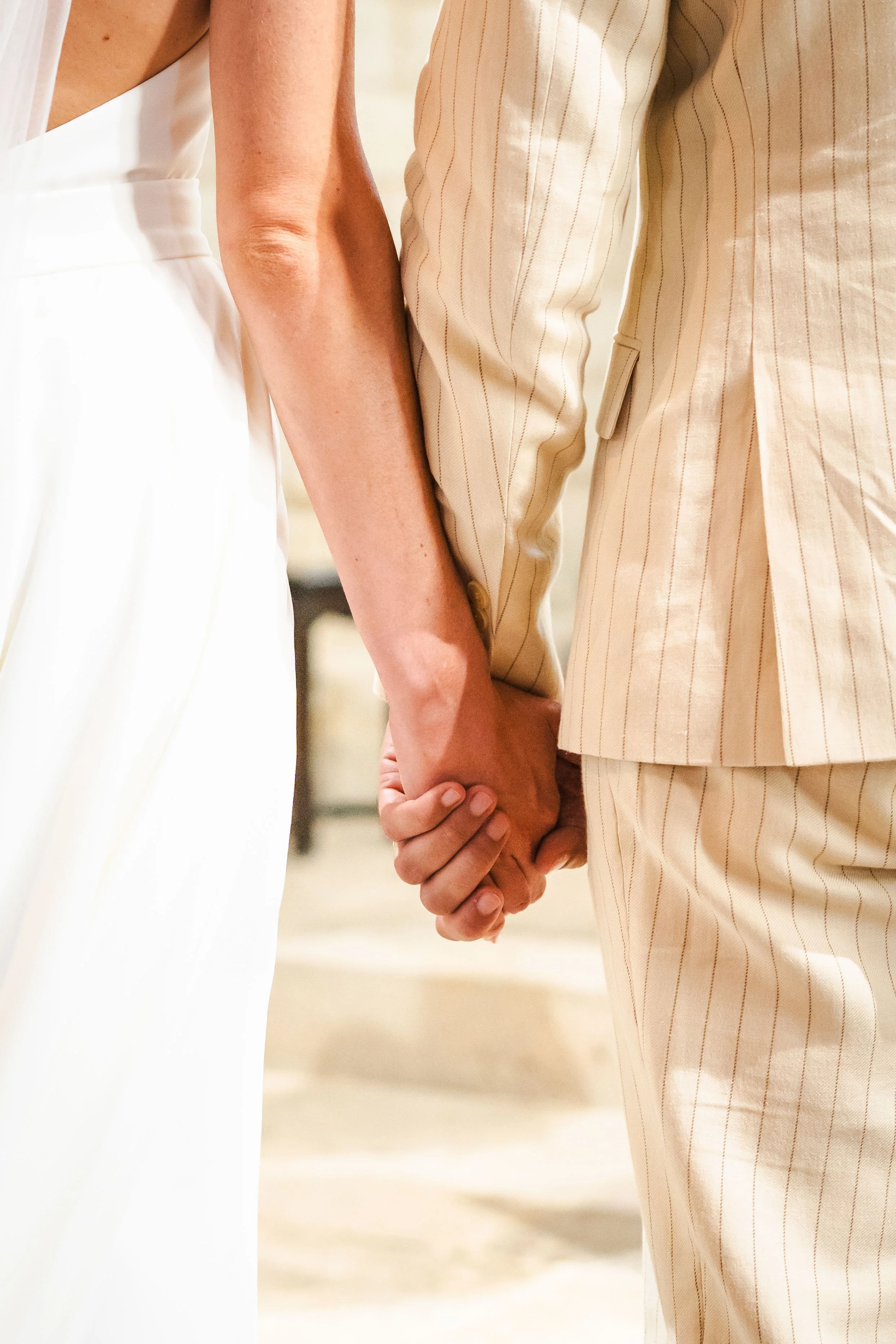 Une personne portant une robe blanche et une autre portant un costume beige à fines rayures, qui se tiennent la main.