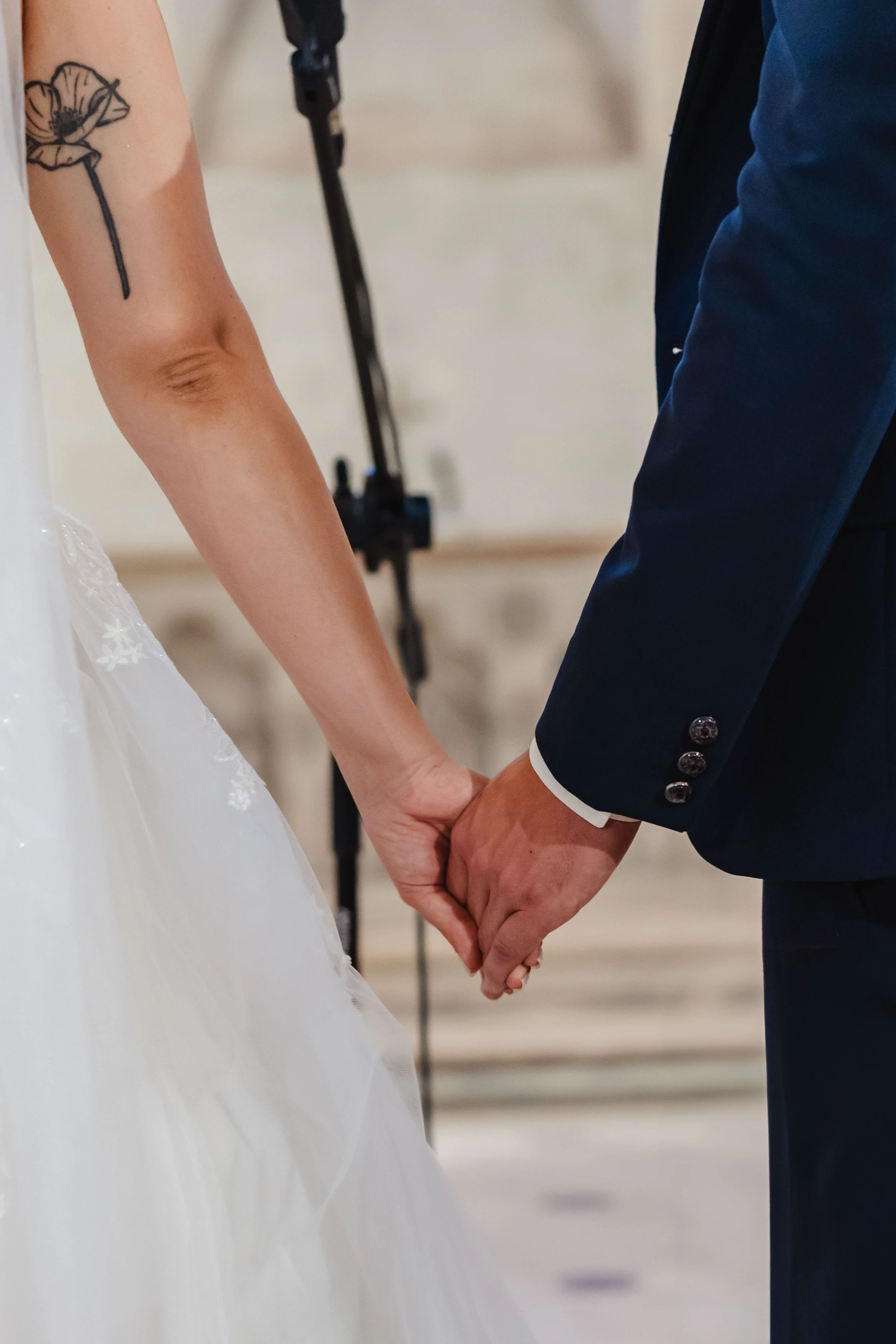 Un couple qui se tient par la main lors de leur cérémonie de mariage, la femme porte une robe de mariée blanche et un tatouage d'une fleur sur le bras, et l'homme porte un costume bleu