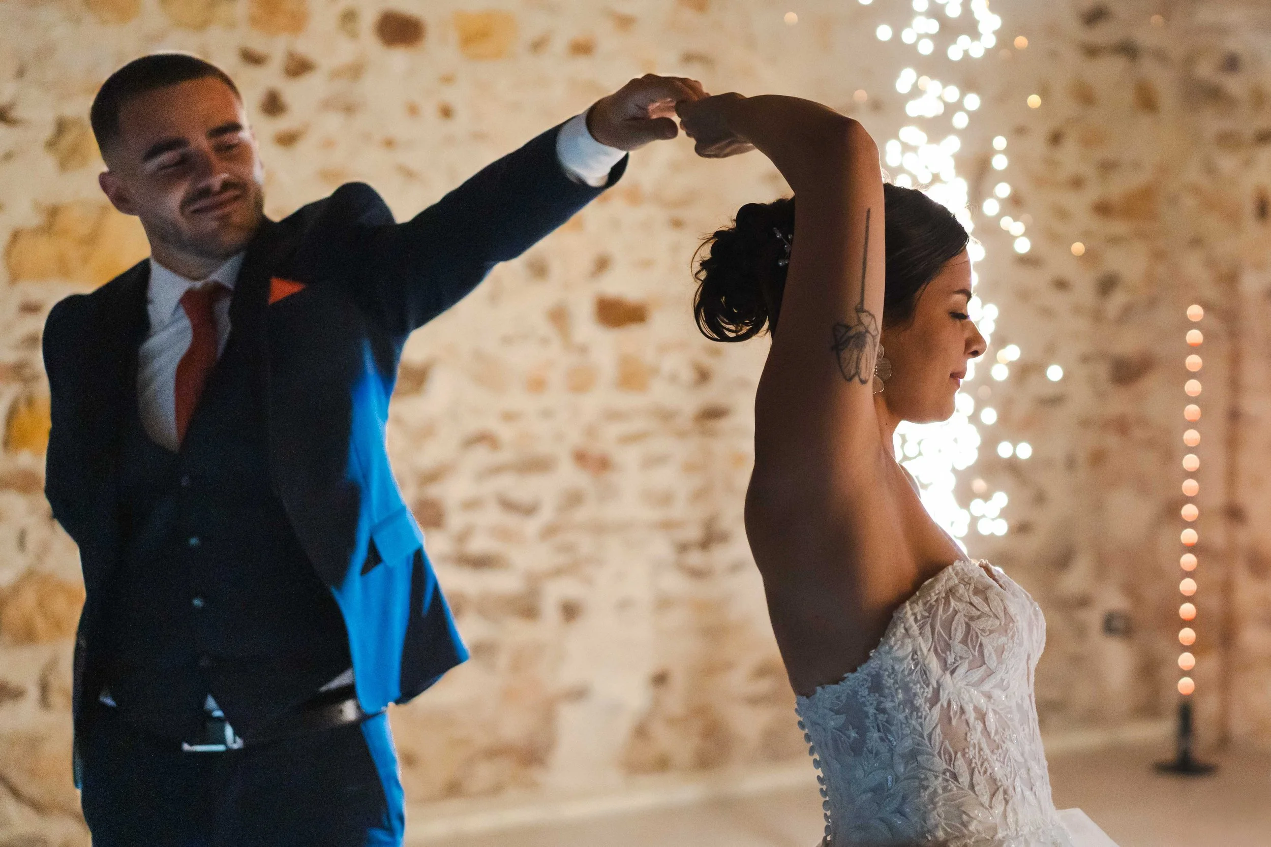 Un couple danse lors de leur mariage, la femme porte une robe de mariée sans manches et un tatouage de papillon sur le bras, l'homme porte un costume sombre avec une chemise blanche et une cravate rouge, ambiance chaleureuse avec des lumières d'ambia