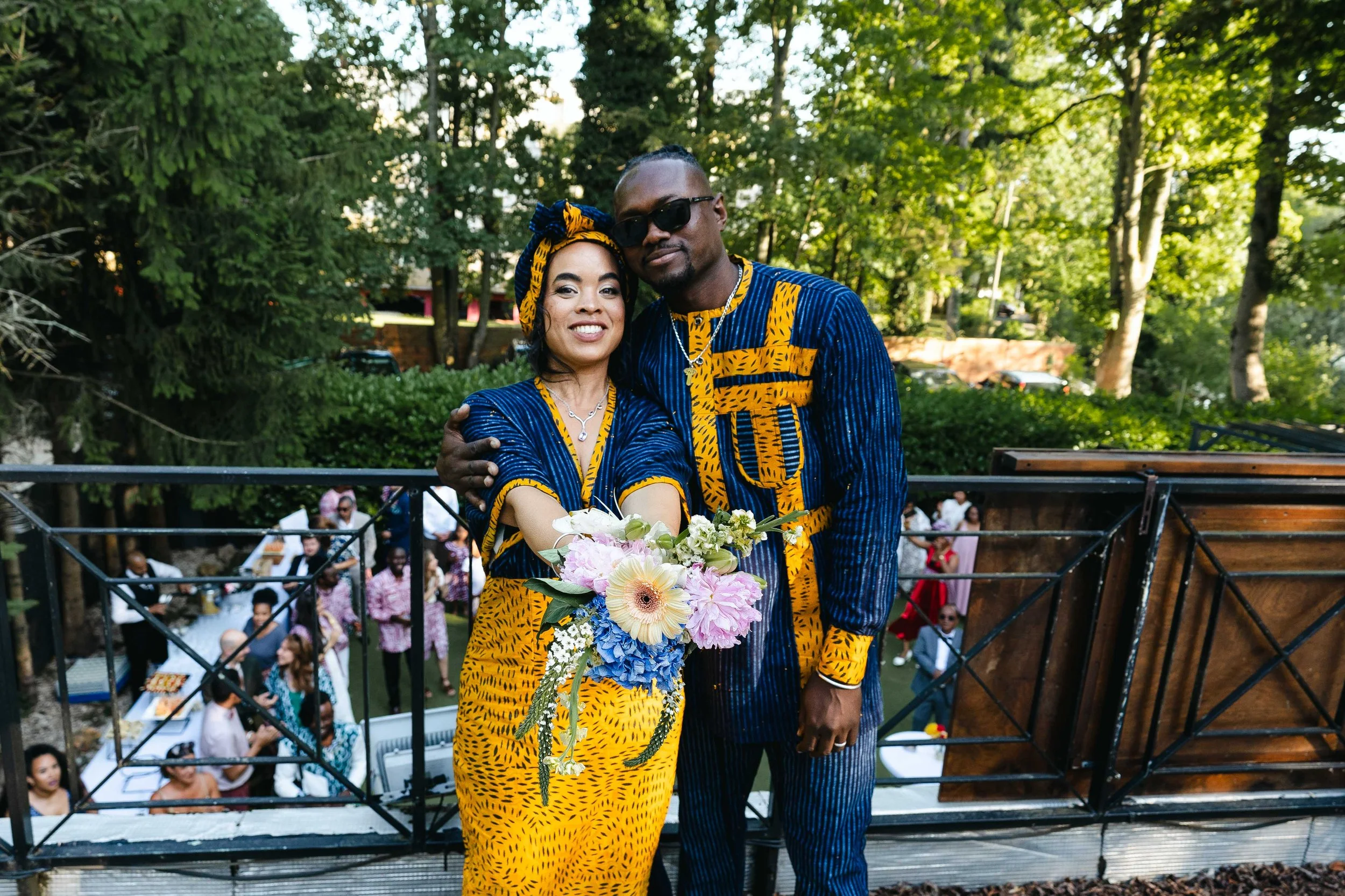 Un couple habillé en vêtements colorés et assortis, posant pour une photo lors d'une célébration en plein air, pour le lancer du bouquet de la mariée, avec un groupe de personnes en arrière-plan dans un parc verdoyant.