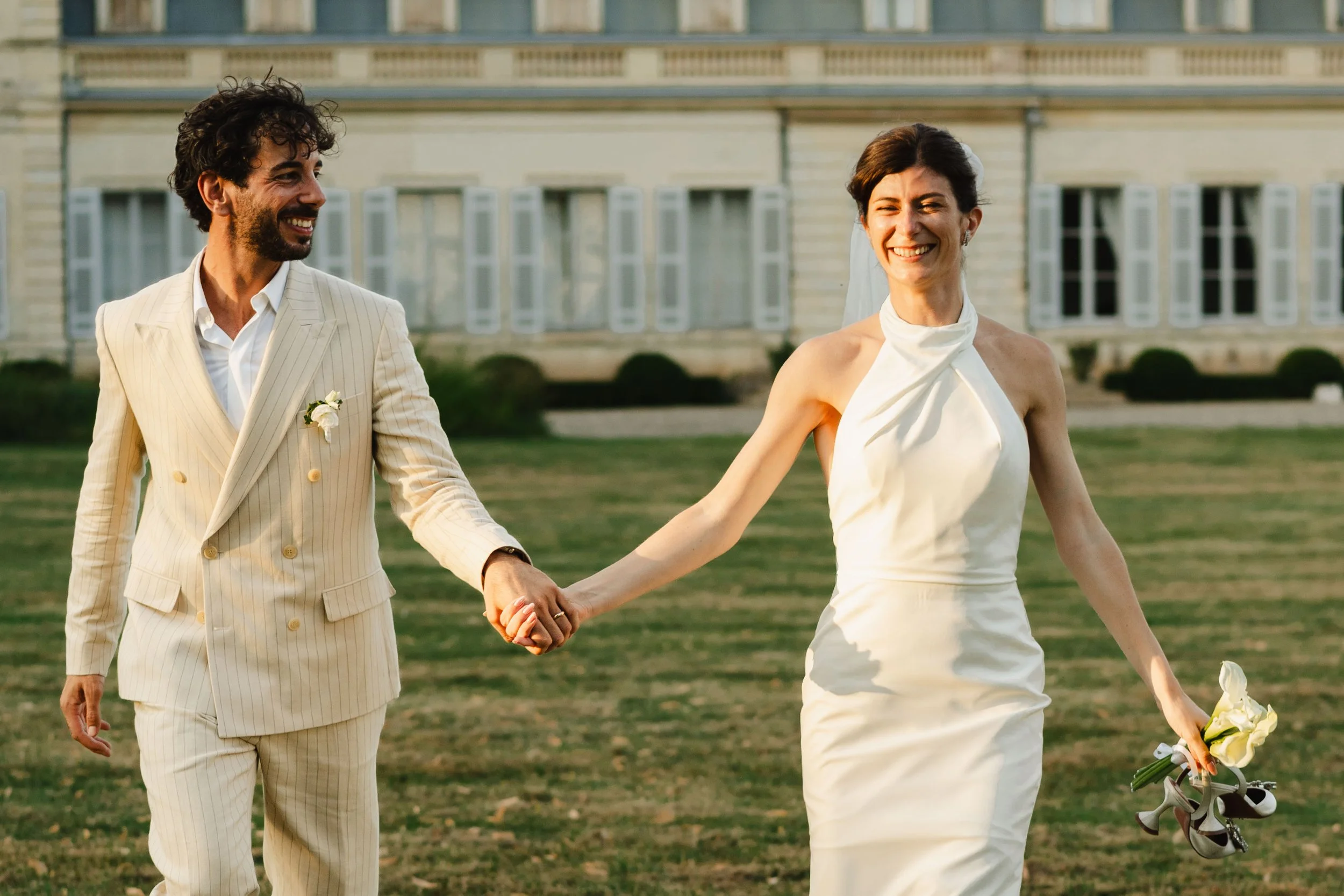 Un couple de mariage souriant marchant main dans la main dans un jardin, la femme porte une robe blanche et tient des chaussures, le homme porte un costume rayé clair.