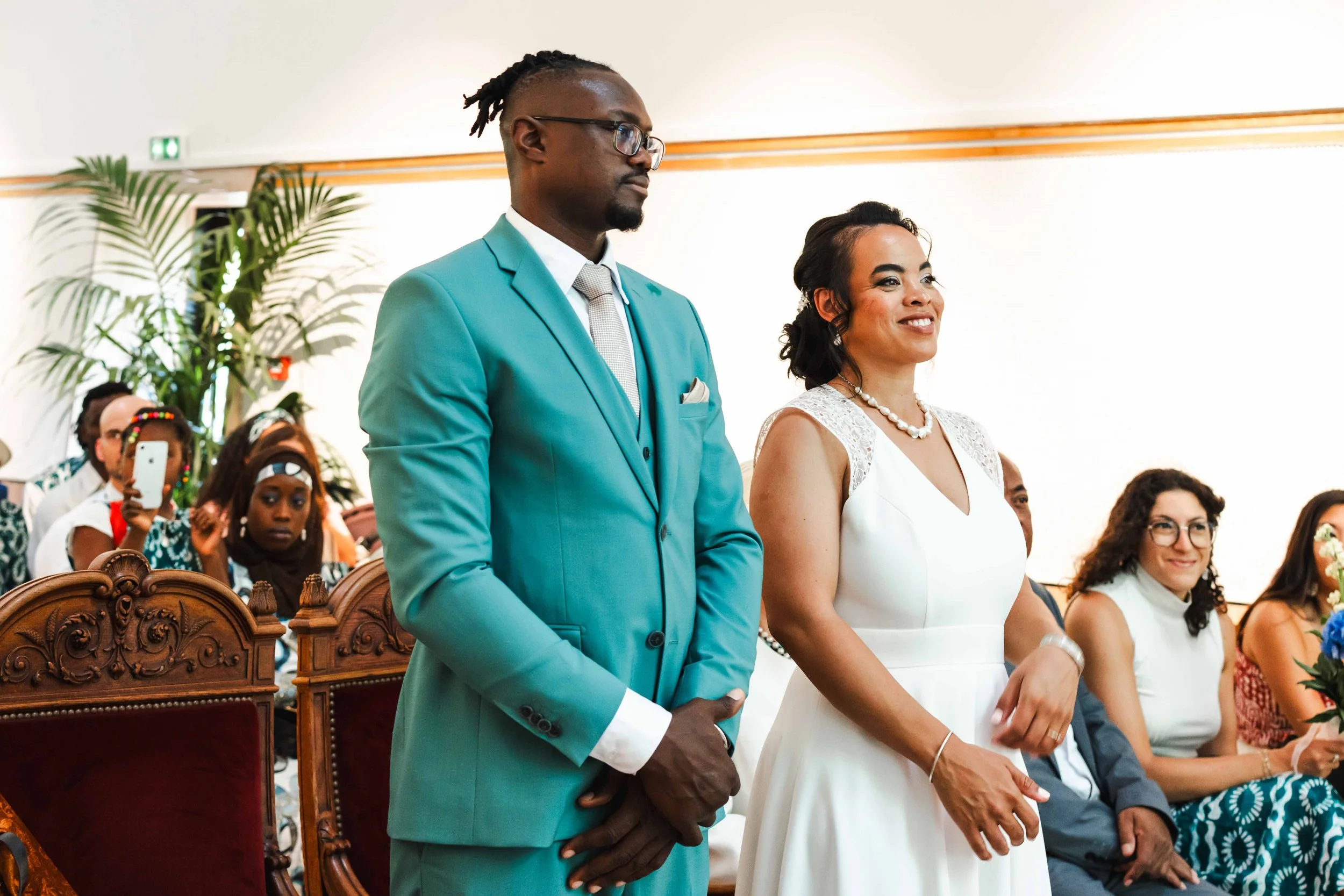 Un couple lors d'une cérémonie, la femme porte une robe blanche et le homme un costume turquoise, entourés d'invités assis.