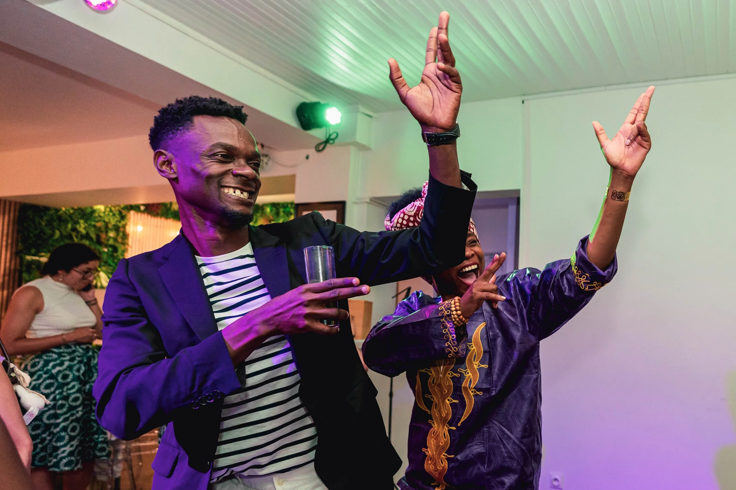 Deux personnes souriantes dans une tenue colorée, en train de danser ou d'agiter les bras lors d'une fête ou d'une célébration intérieure, avec une ambiance festive et éclairage coloré.
