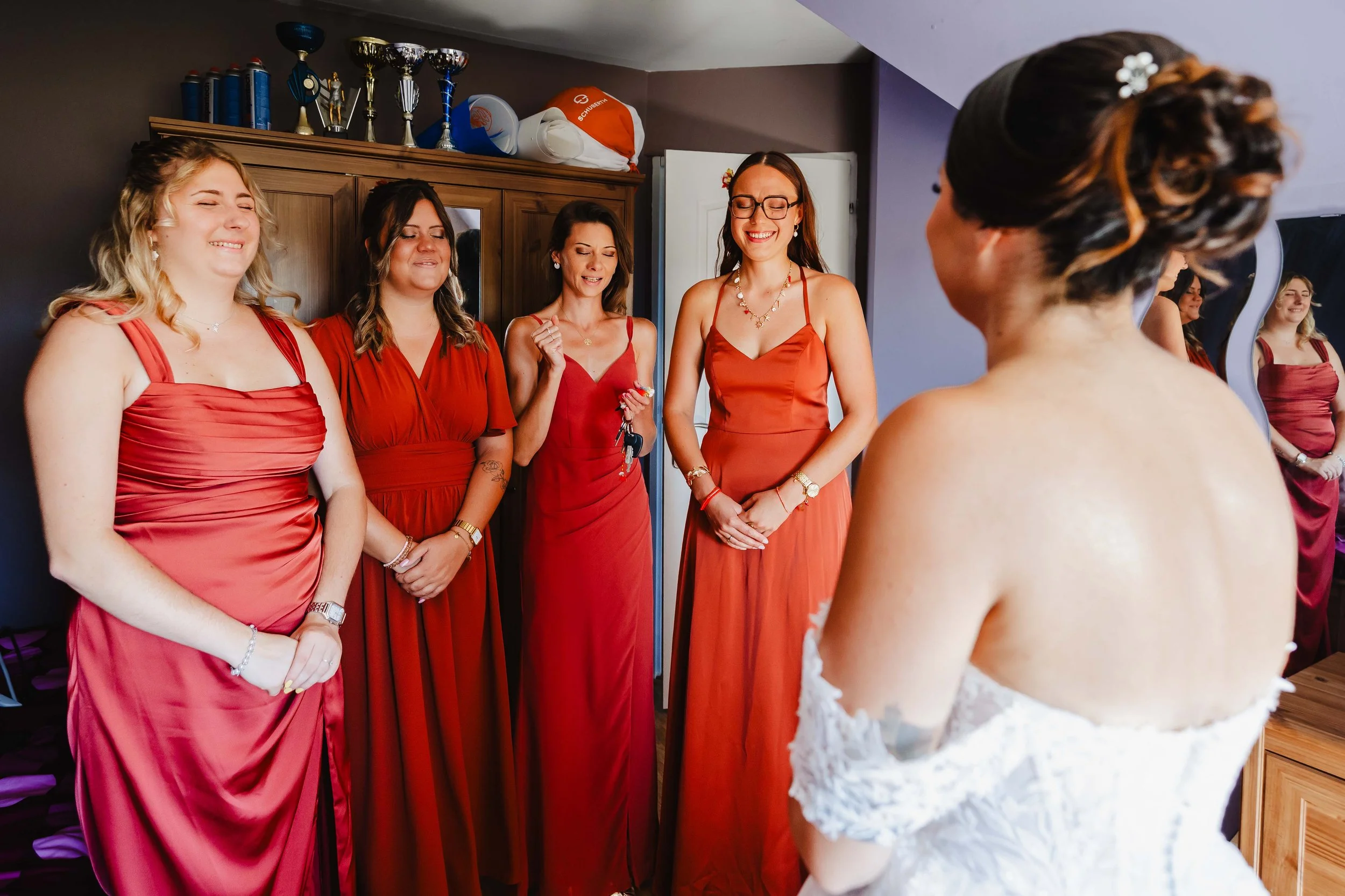 Fille en robe de mariée en train de voir ses amies en robes rouges lors d'une réunion pré-mariage dans une pièce avec armoire et trophées au mur.