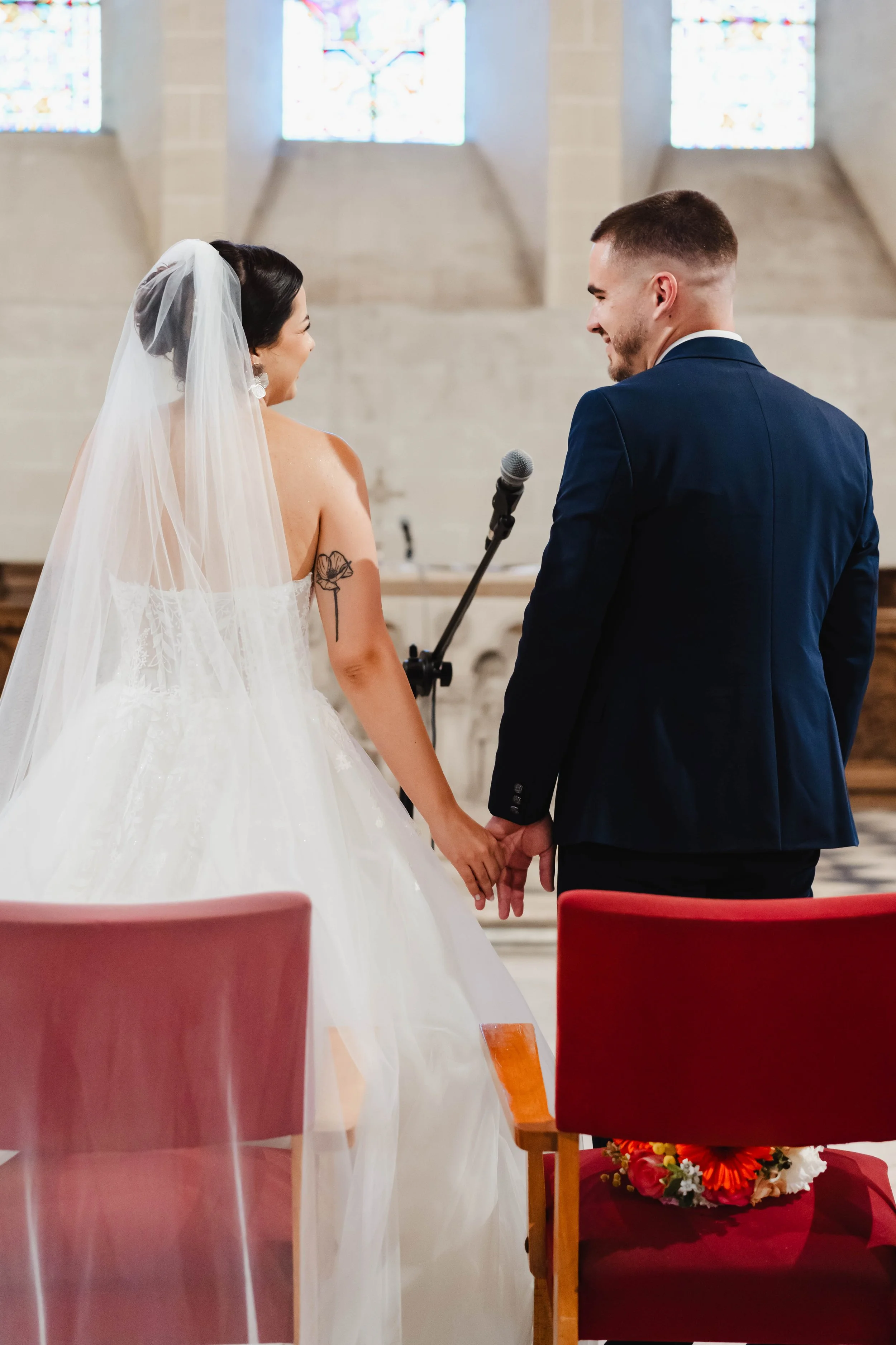 Un couple de mariés lors de leur mariage religieux dans l'église de Saint Pierre Les Neymours, tenant la main avec des chaises rouges en avant et des vitraux en arrière-plan.
