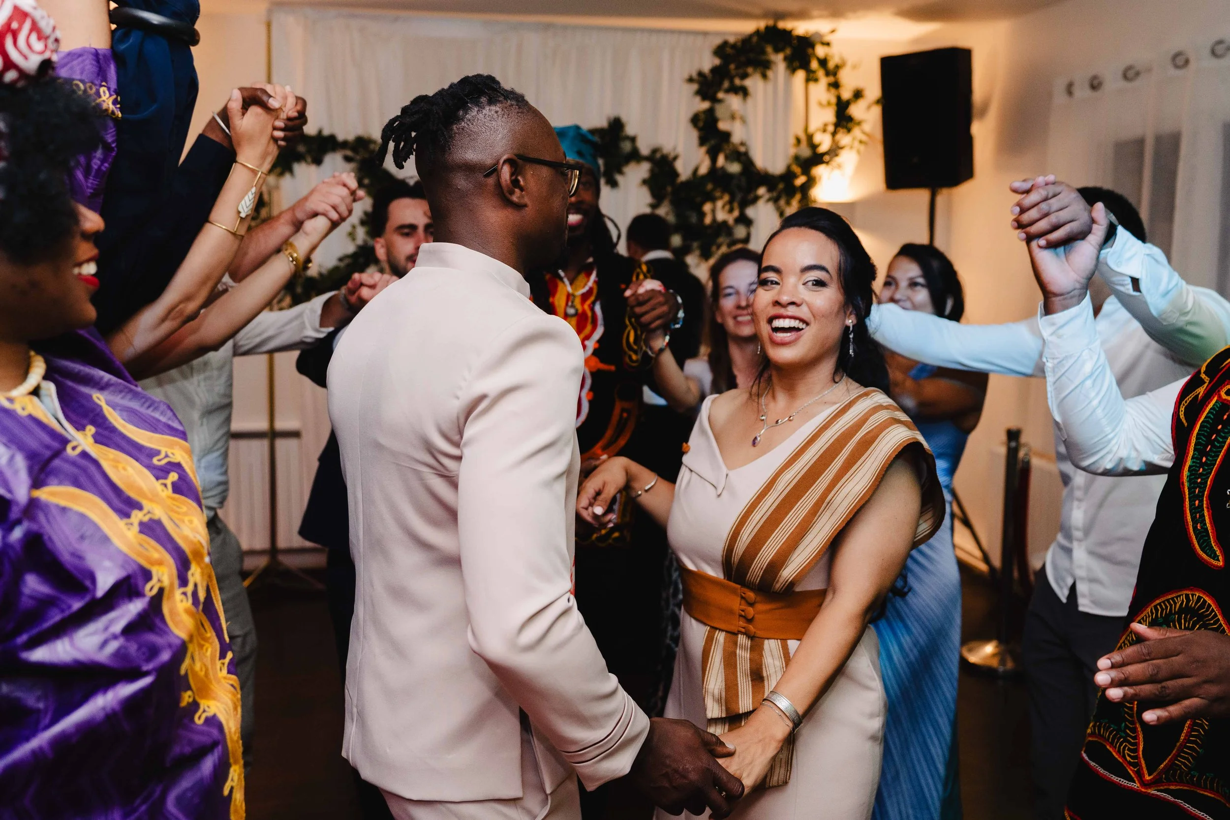 Groupe de personnes dansant et souriant lors d'une célébration ou d'une fête dans une salle décorée, avec un couple au centre tenant la main et regardant l'un l'autre.