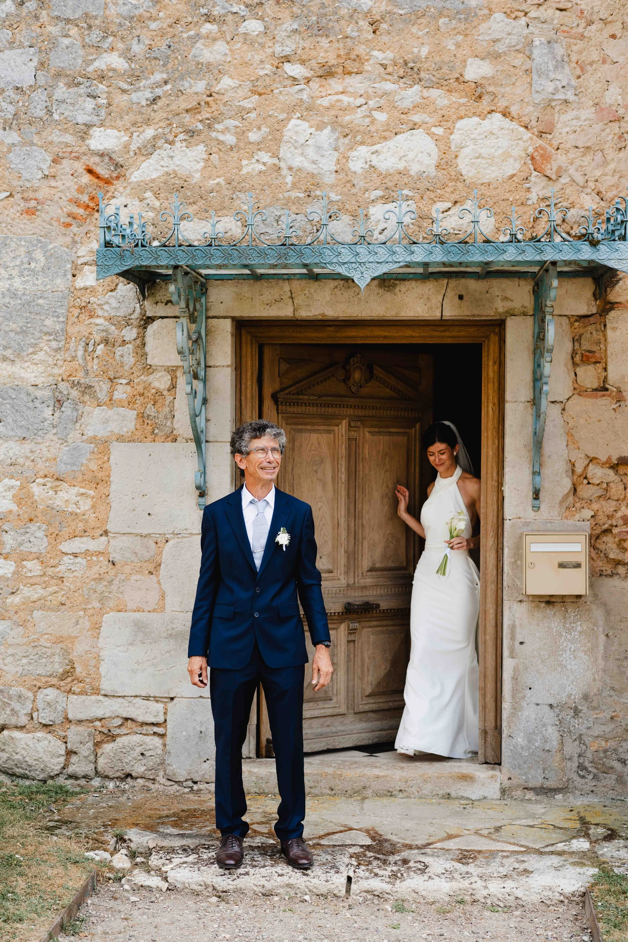 Un homme en costume avec une boutonnière blanche se tenant devant la porte d'une vieille maison en pierre, avec une jeune femme en robe de mariée à l'intérieur qui tient un bouquet.