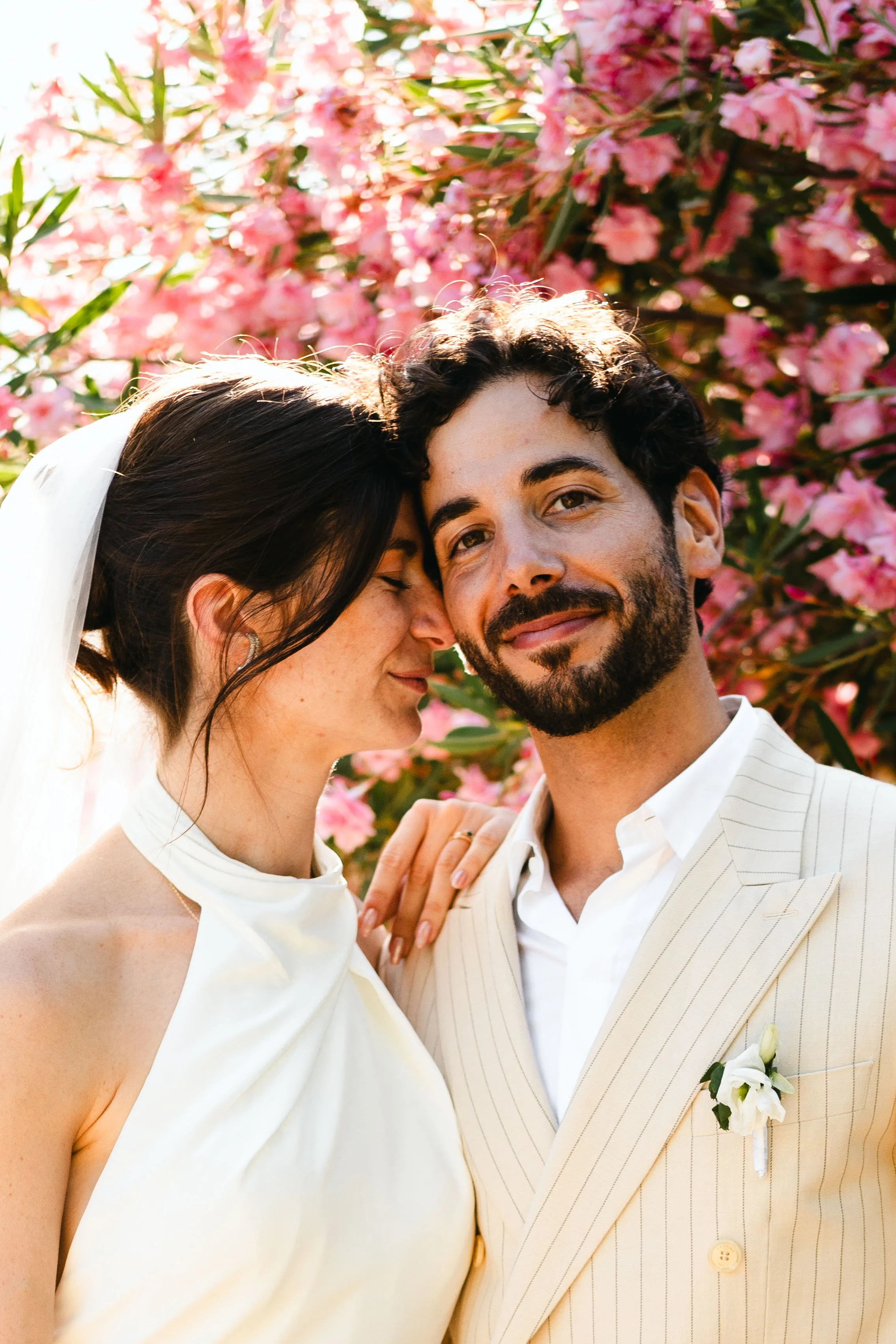 Un couple de mariés pose près d'un arbre en fleurs roses. La femme porte une robe de mariage blanche et un voile, et l'homme porte un costume beige rayé avec une boutonnière blanche. Ils semblent heureux et sont très proches.
