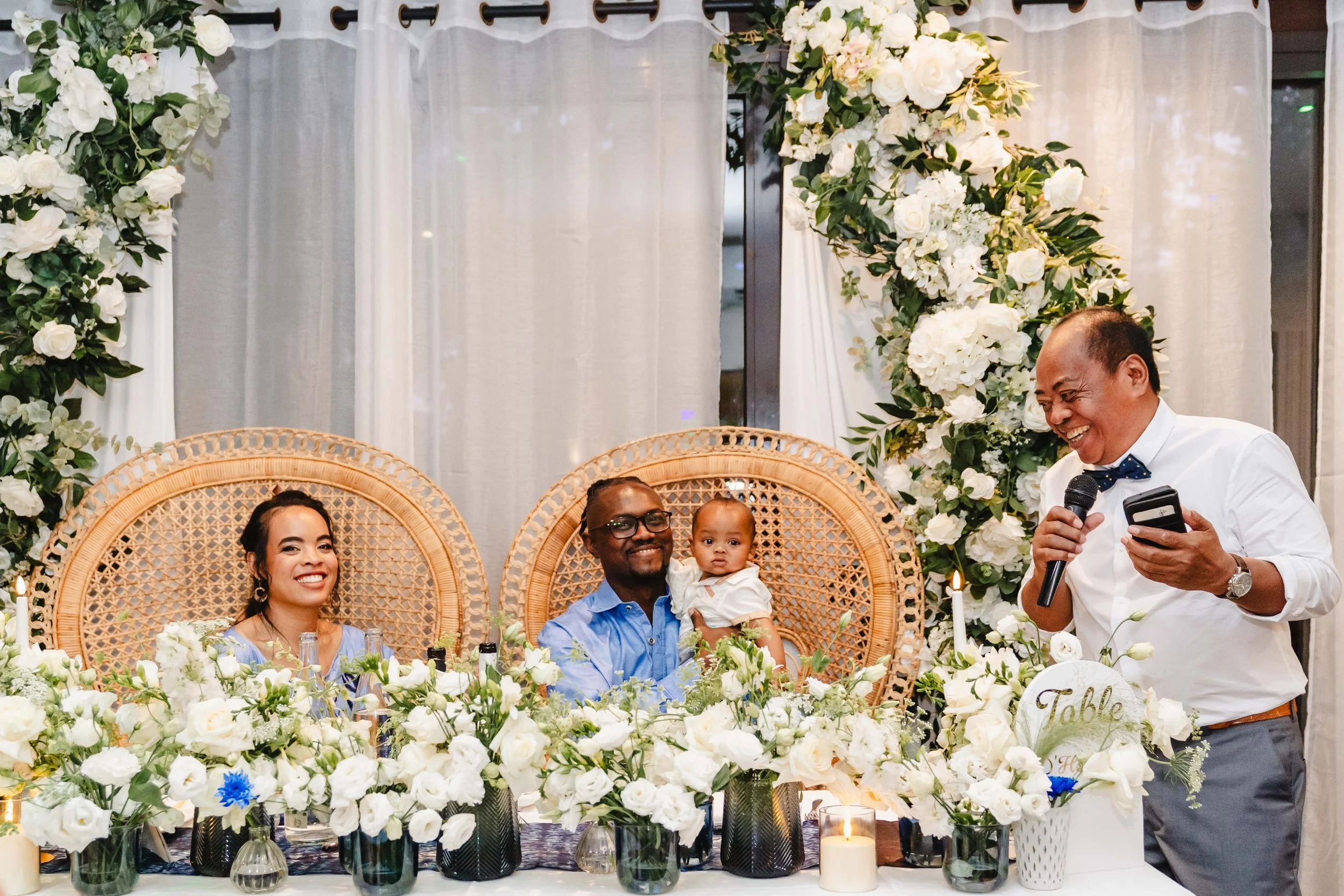 Une famille lors d'une célébration, assise à une table décorée avec des fleurs blanches, un homme tenant un micro et un téléphone, souriant, avec deux autres personnes, un homme et une femme, ainsi qu'un bébé. L'arrière-plan est décoré de fleurs blan