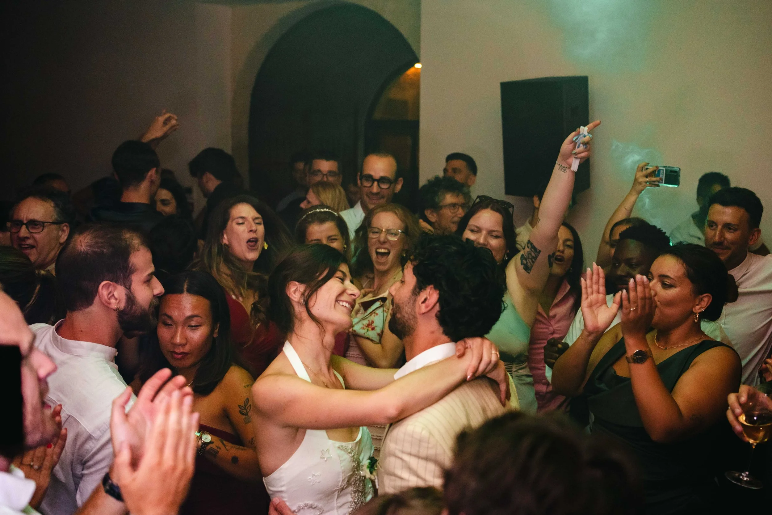 Un groupe de personnes souriantes et joyeuses à une fête ou un mariage, dansant et prenant des photos, avec un couple en particulier qui s'embrasse au centre dans la salle de réception du chateau Saint Denis.