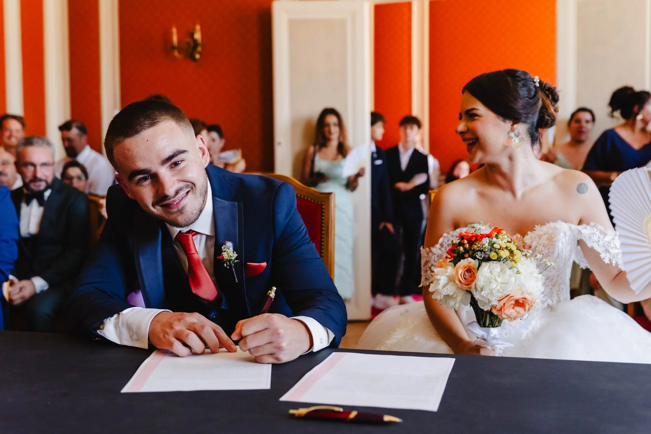 Un couple de mariés assis à une table lors d'un mariage, l'homme signe un document et la femme, tenant un bouquet de fleurs, lui sourit dans une salle ornée de murs rouge foncé et de nombreux invités en arrière-plan.