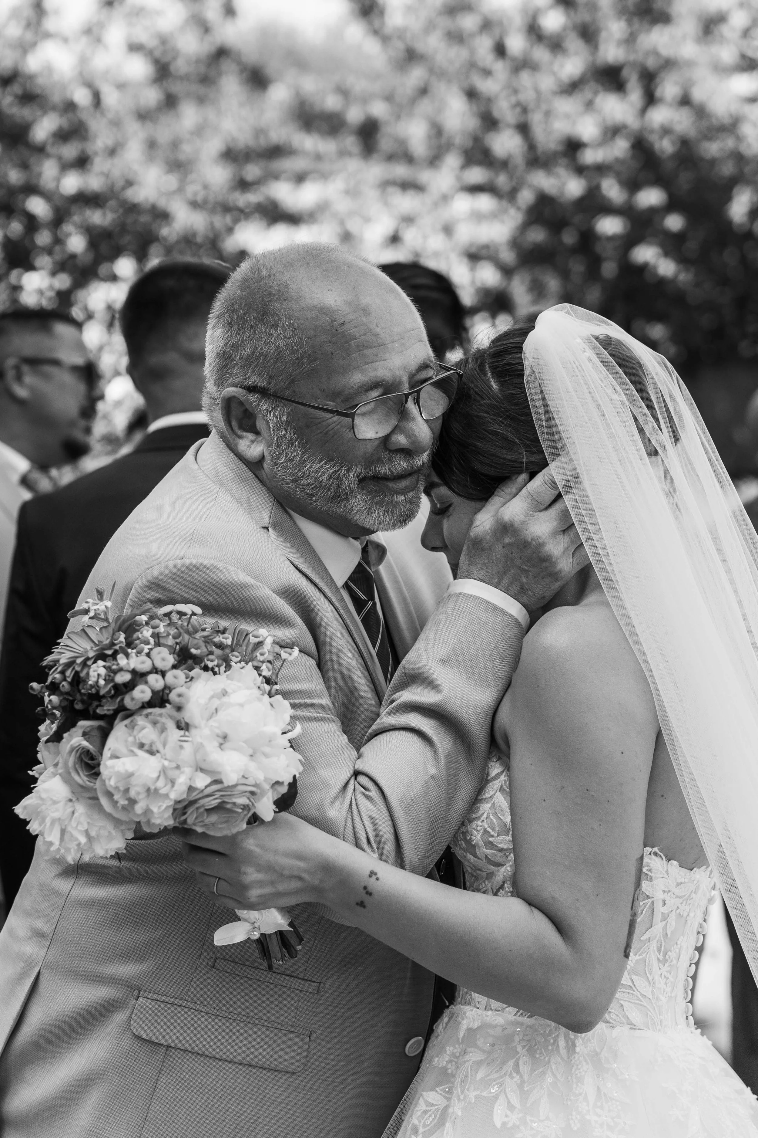 Un homme plus âgé embrasse une jeune femme en robe de mariée lors d'une cérémonie de mariage, avec un bouquet de fleurs à la main, dans un cadre extérieur.