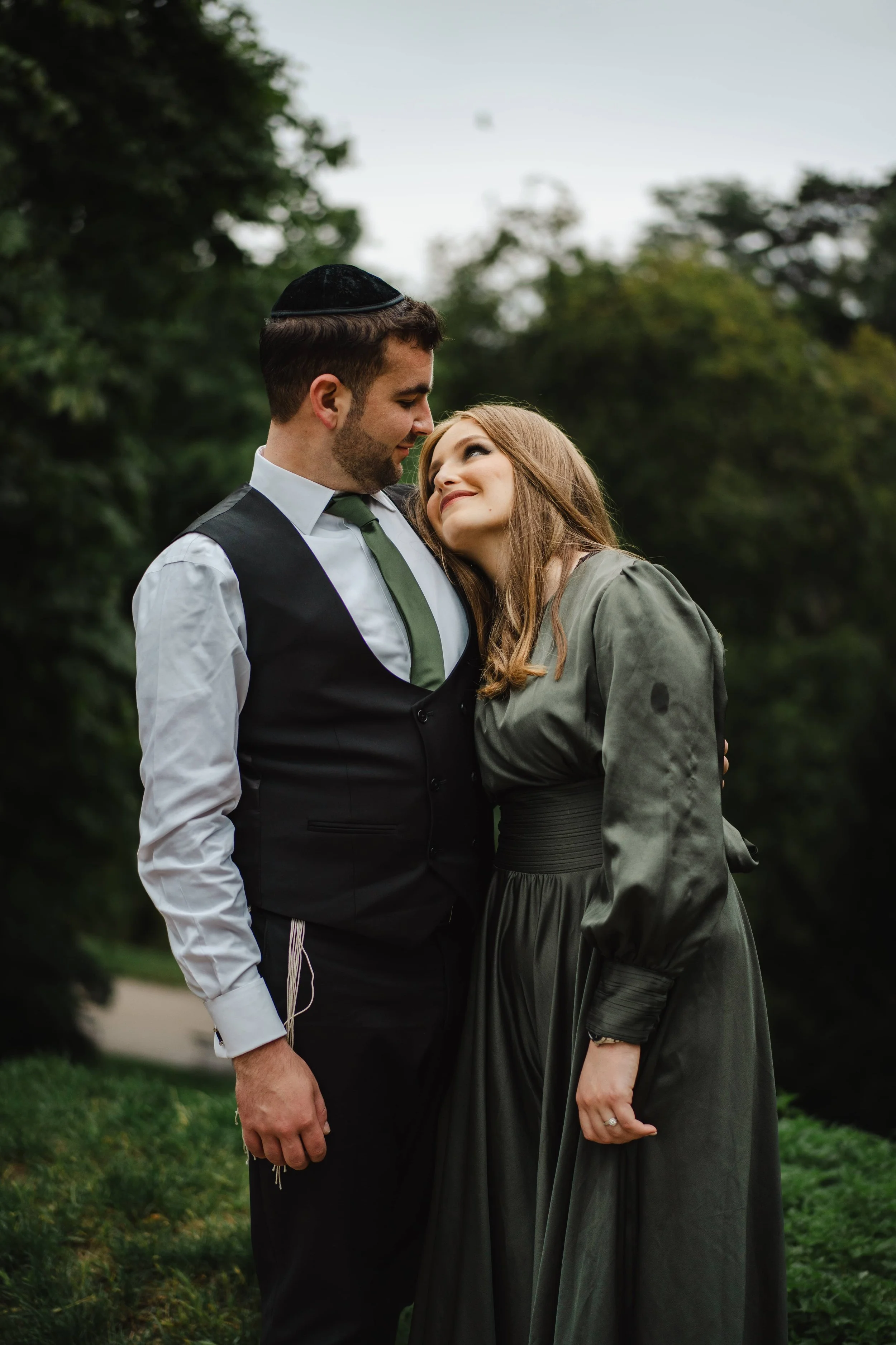 Un couple bien habillé, un homme portant une kippa, et une femme habillée d'une robe longue, se regardent affectueusement dans le parc des Buttes Chaumont.