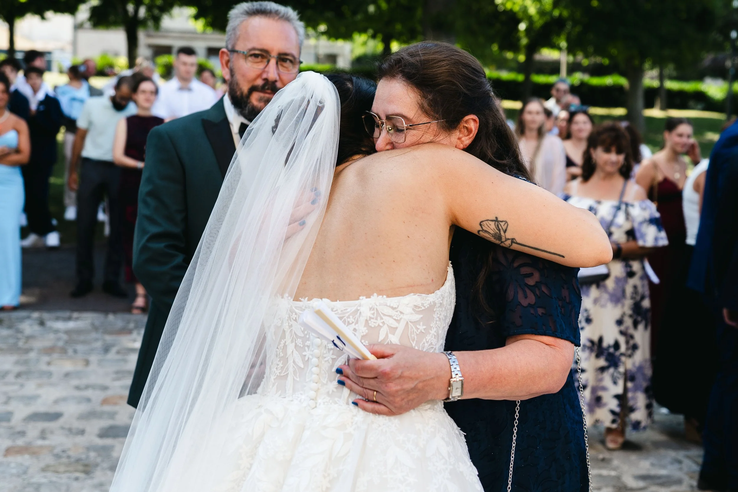 Une mariée en robe blanche embrasse une femme lors d'un mariage en plein air, d'autres invités en arrière-plan