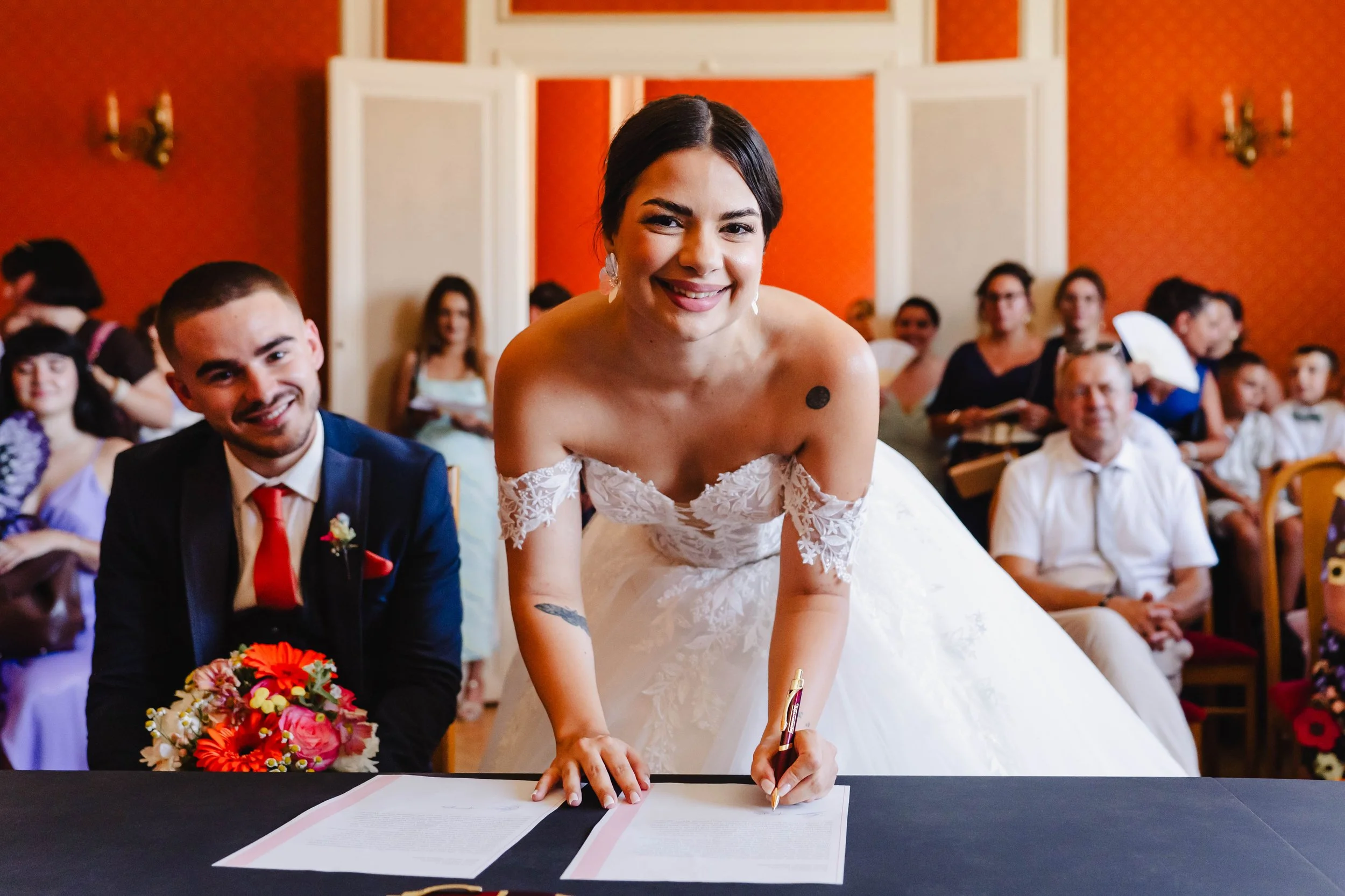 Une femme en robe de mariée signe un contrat lors d'une cérémonie de mariage, entourée de personnes assises dans une salle ornée de murs rouge vif, dans une ambiance joyeuse.