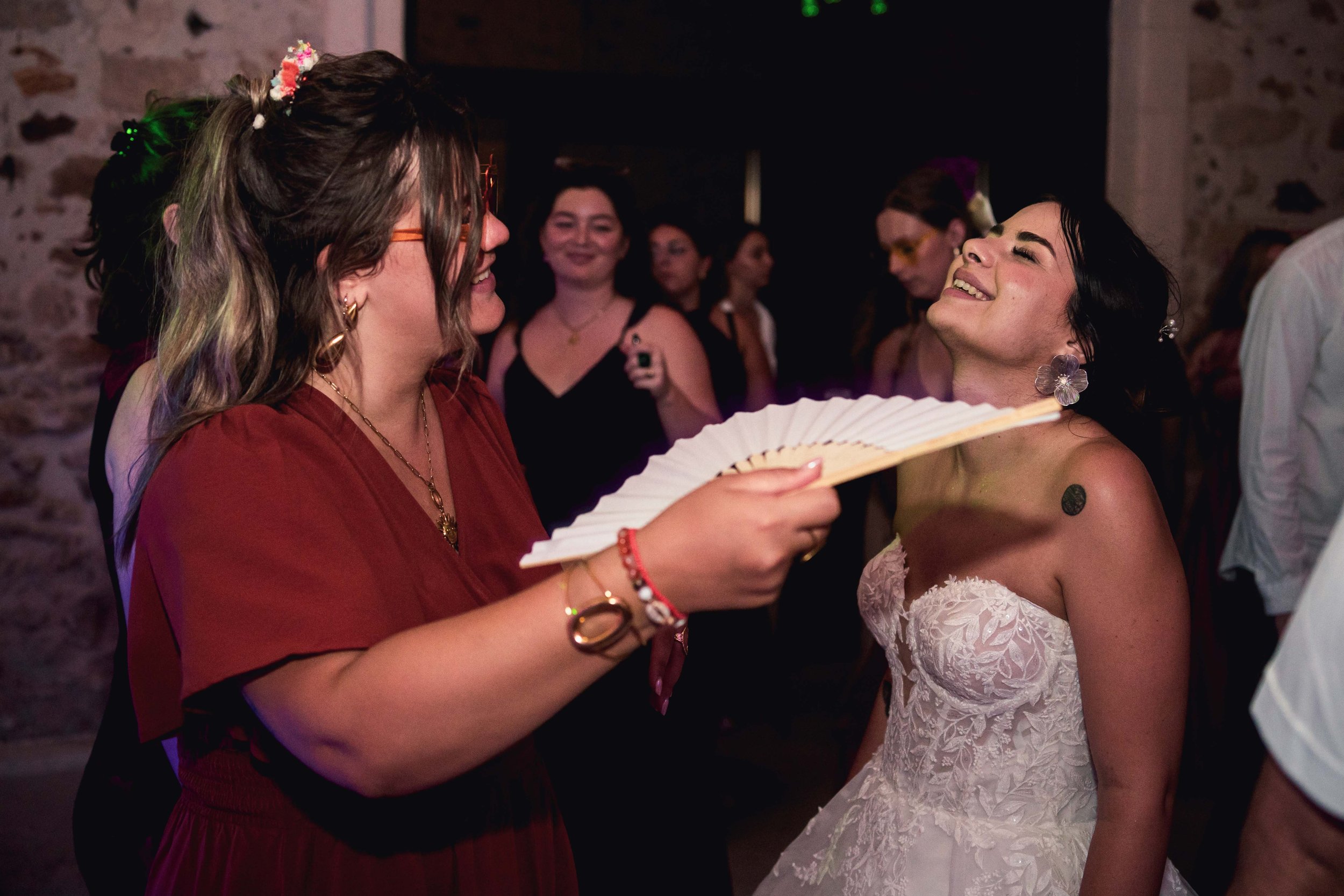 Deux femmes à une fête, l'une portant une robe de mariage, l'autre une tenue rouge, jouent avec un éventail en papier blanc, partageant un moment joyeux parmi d'autres invités dans une ambiance festive.