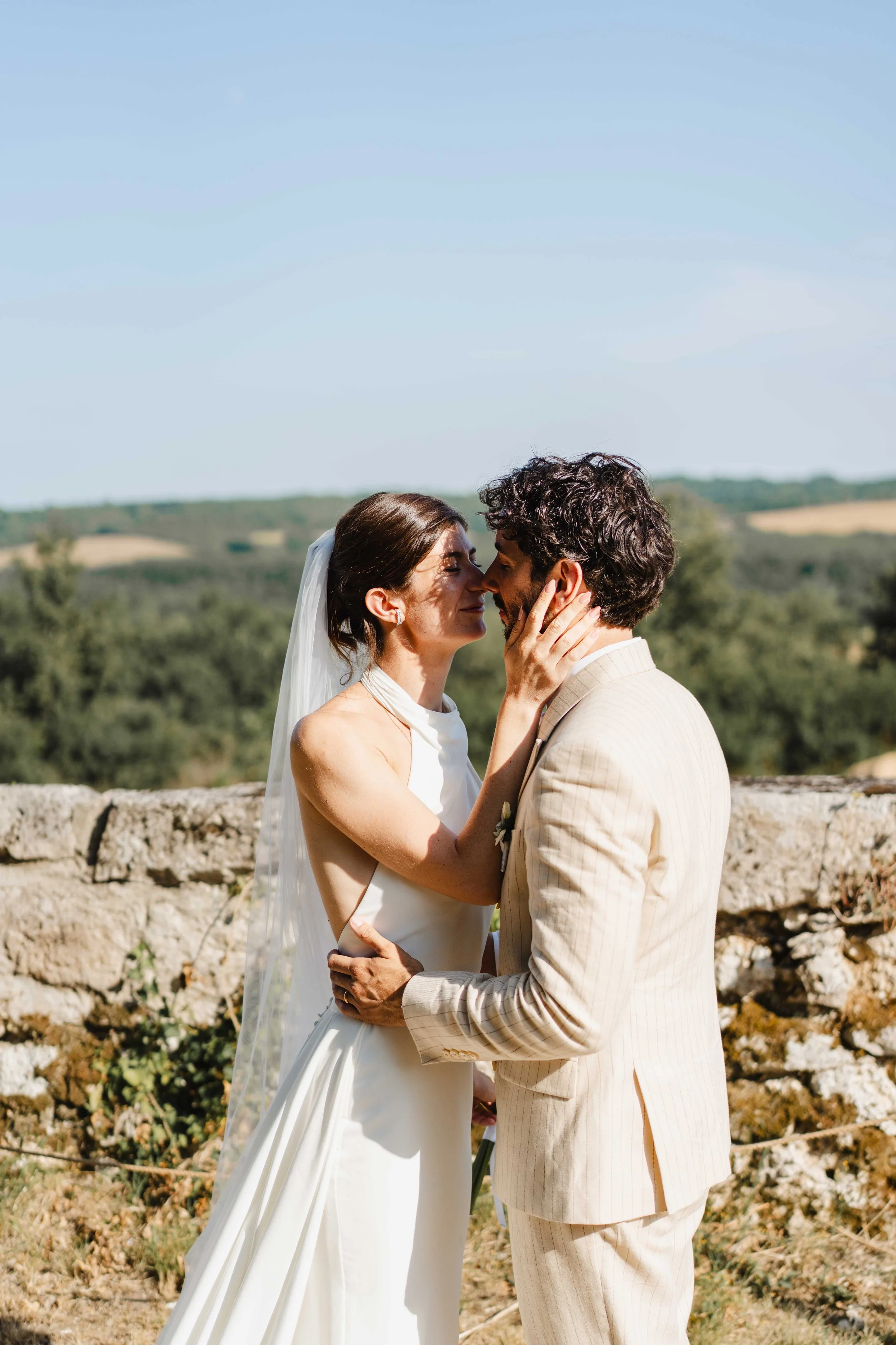 Un couple en robe de mariage et costume en train de s'embrasser lors d'une cérémonie en plein air avec un paysage verdoyant en arrière-plan.