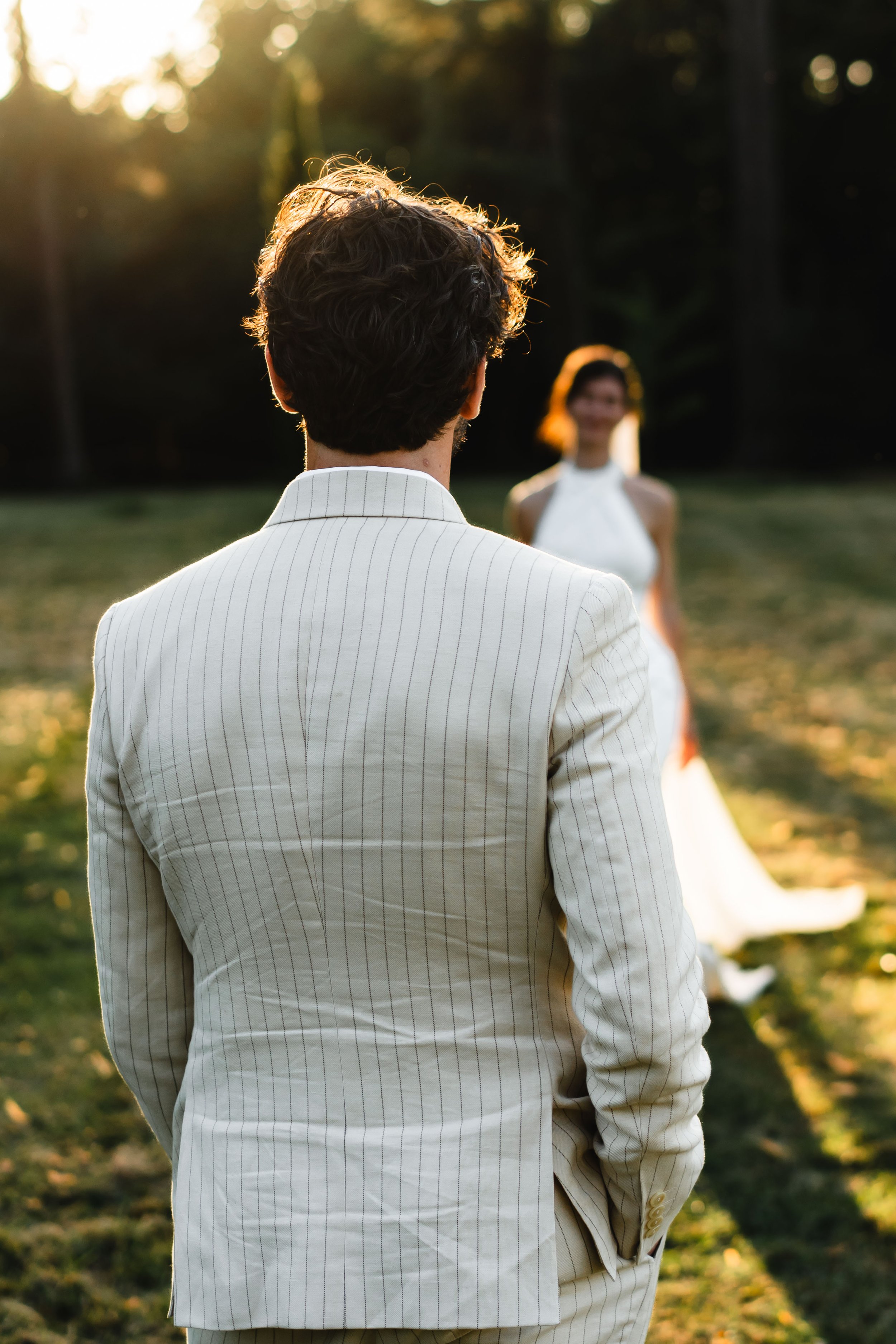 Un homme en costume blanc regarde une femme en robe blanche lors d'une cérémonie dans un extérieur en plein air, avec les rayons du soleil couchant en arrière-plan.