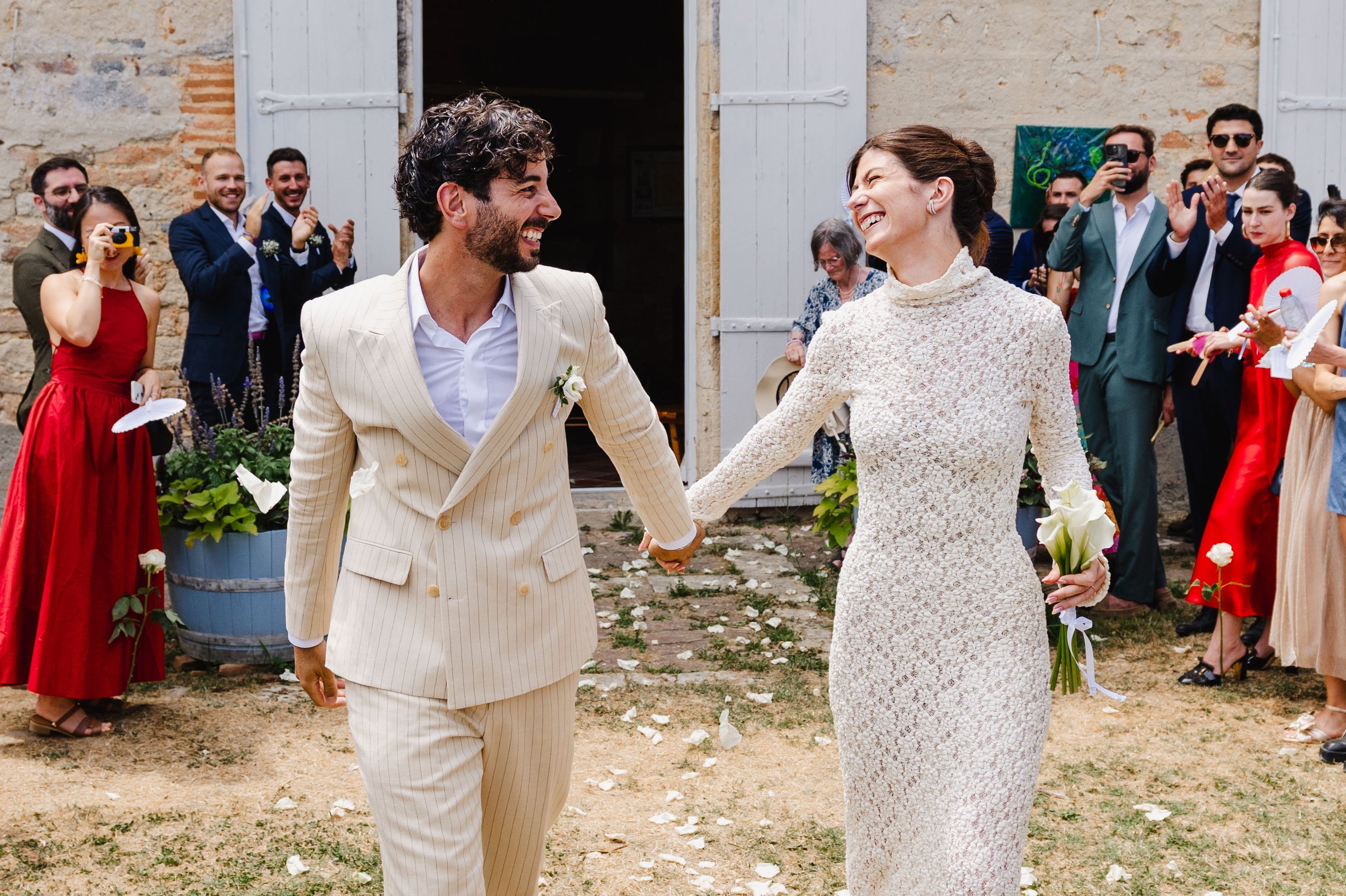 Un couple de mariés tenant la main, souriants, lors de leur mariage en plein air, entourés d'amis et de famille applaudissant, avec des décorations florales et un bâtiment en pierre, la mairie de Moirax,  en arrière-plan.