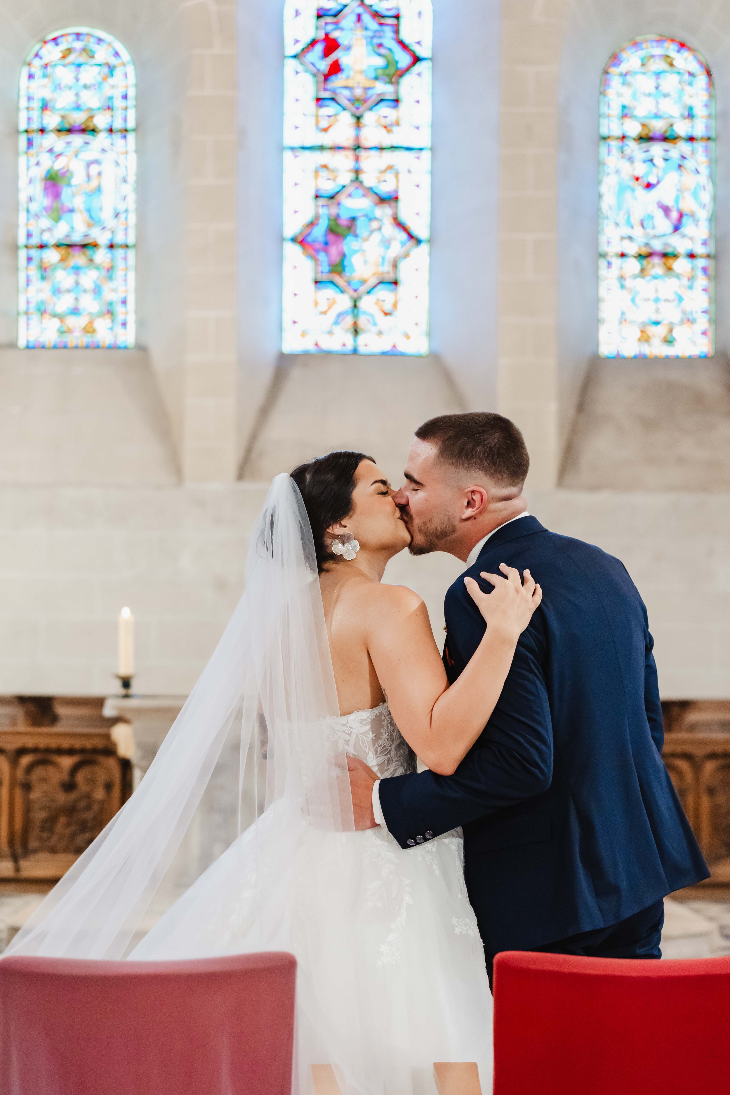 Un couple de mariés s'embrassant dans l'église de Saint Pierre Les Neymours avec des vitraux colorés à l'arrière-plan.