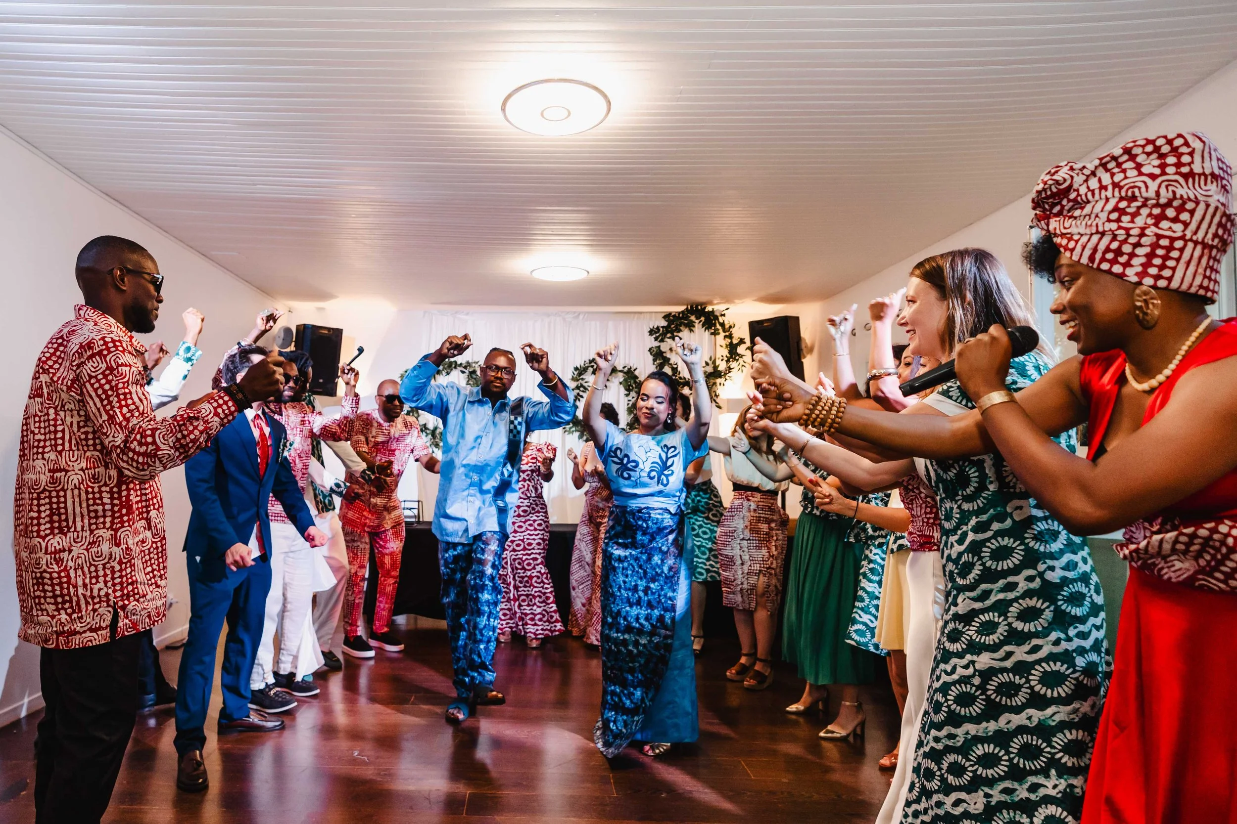 Groupe de personnes dansant et chantant lors d'une célébration, vêtues de tenues traditionnelles colorées, dans une salle décorée pour un événement festif.