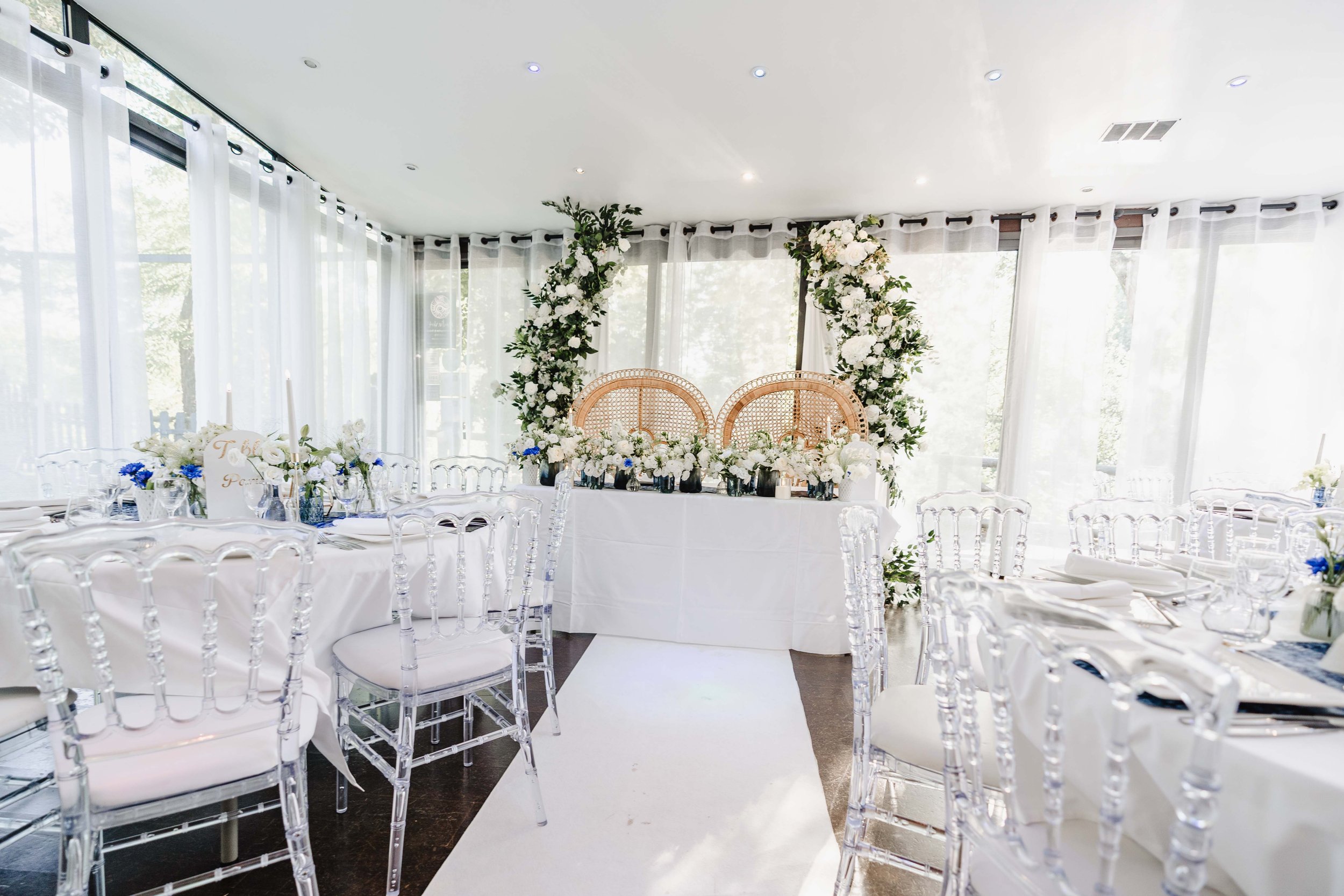 Salle de mariage élégante du moulin vert décorée de fleurs blanches et vertes, avec un arc floral derrière une table d'honneur, entourée de chaises transparentes.
