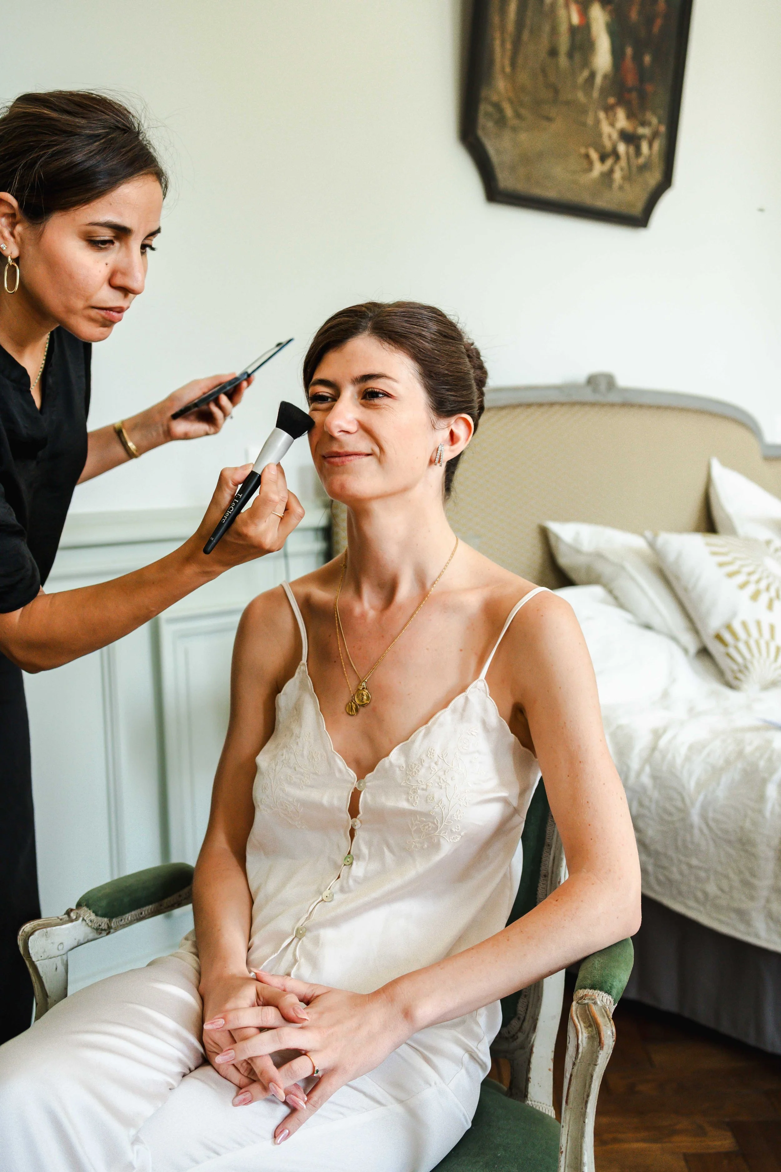 Une femme est assise en train d'être maquillée, avec l'artiste maquilleuse Hannah Ka appliquant du maquillage sur son visage dans une pièce élégante.