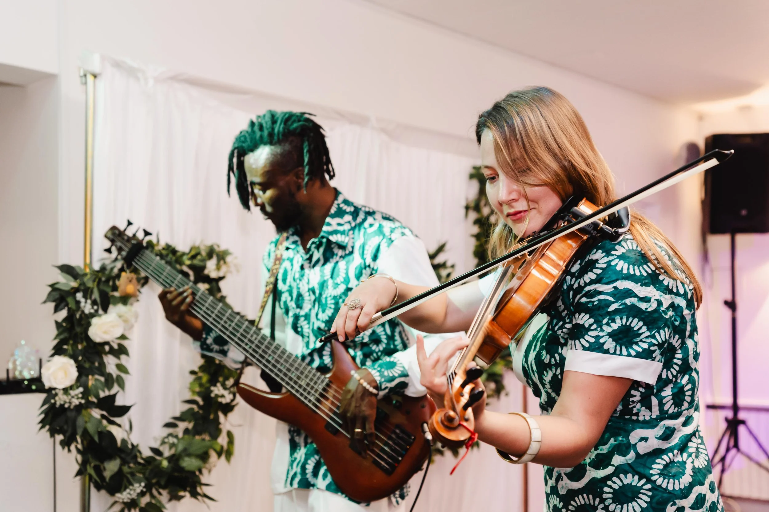 Deux musiciens jouant du violon et de la guitare lors d'une performance dans une ambiance chaleureuse et décorée de fleurs.