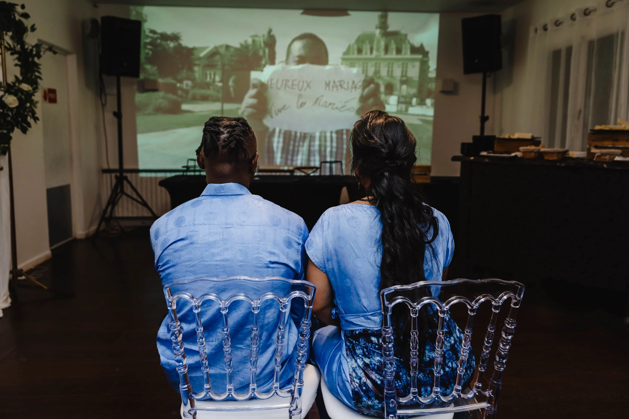 Deux personnes assises sur des chaises transparentes regardant une projection vidéo où une personne tient une pancarte avec l'inscription «  VIVE LES MARIES » en français.