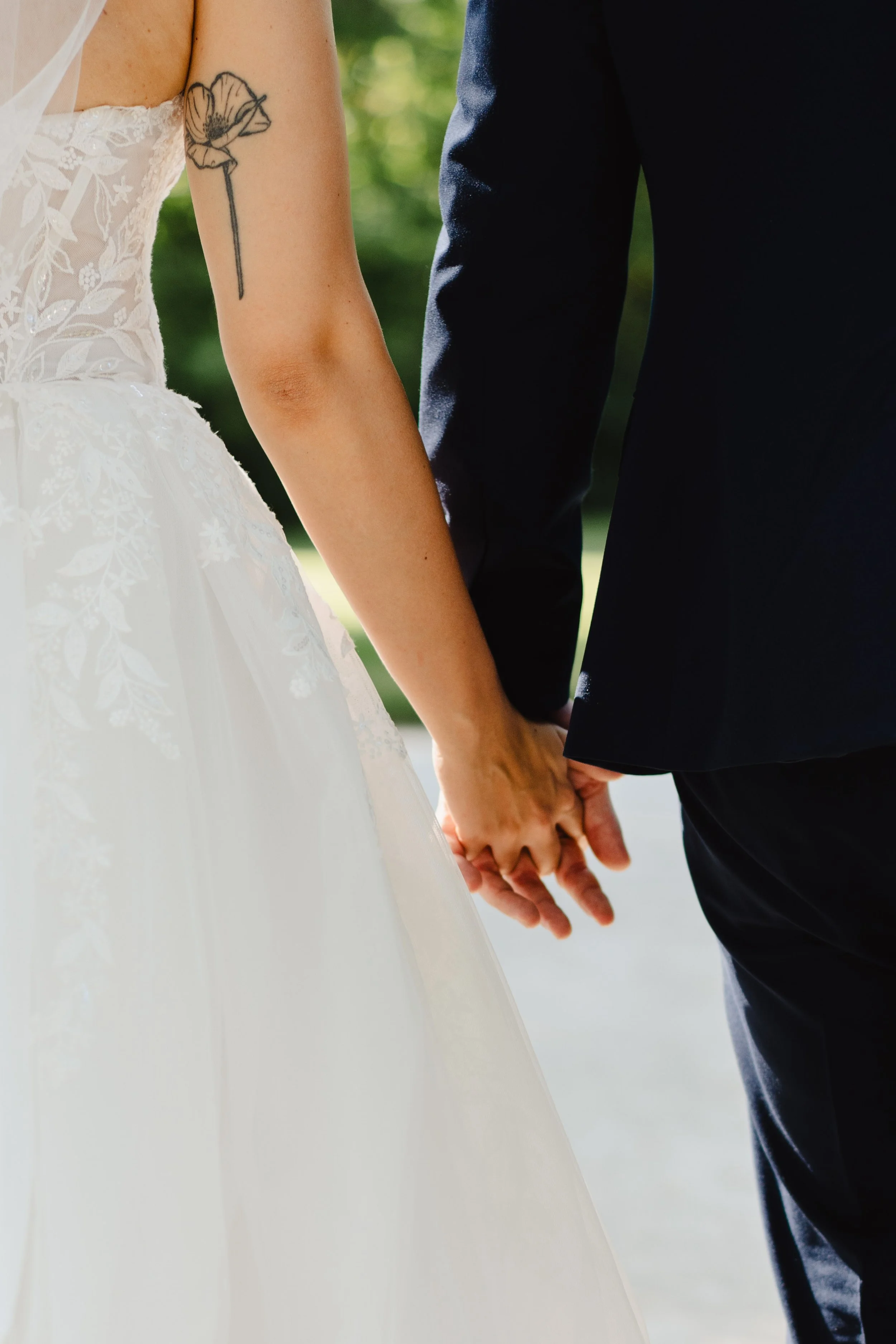 Un couple en costume de mariage se tient la main, vu de derrière, en plein air avec un fond de verdure.