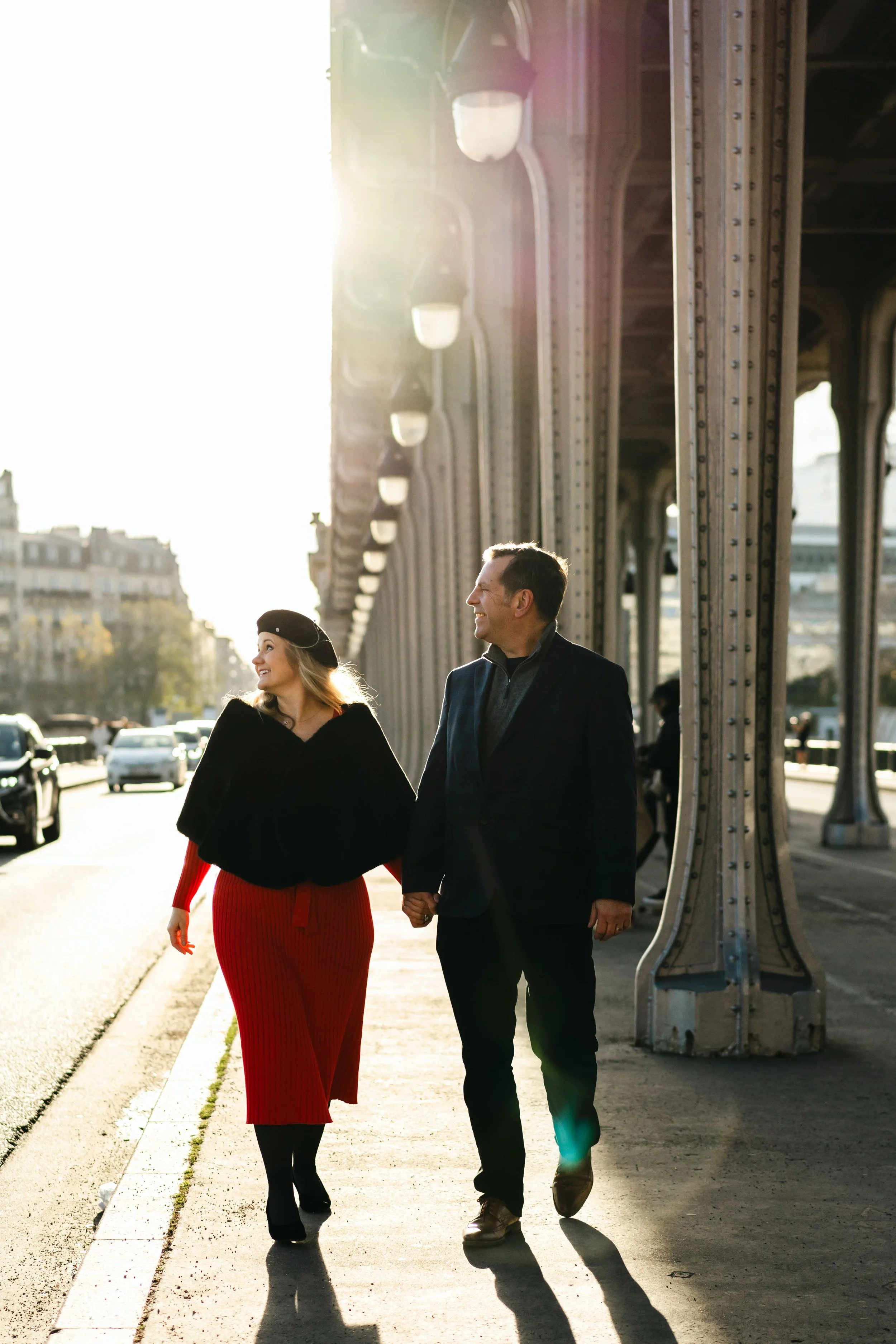 Un couple marche main dans la main sous un pont à Paris en arrière-plan, avec le soleil brillant derrière eux, créant un effet de silhouette et de lumière chaude.