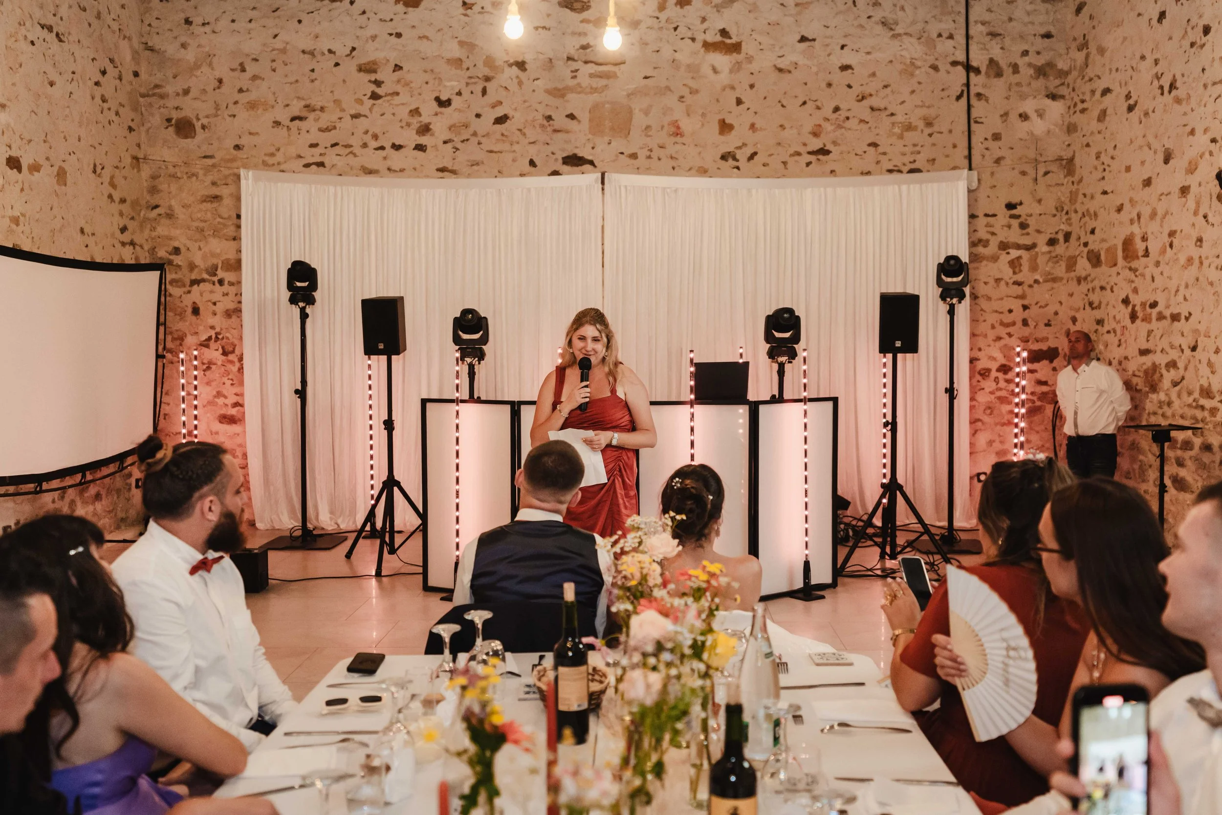 Une femme en robe rouge parle avec un micro lors d'un événement dans une salle décorée avec des murs en pierre et une toile blanche derrière. Des invités sont assis à une table élégamment dressée avec des fleurs, des bouteilles de vin et des appareil