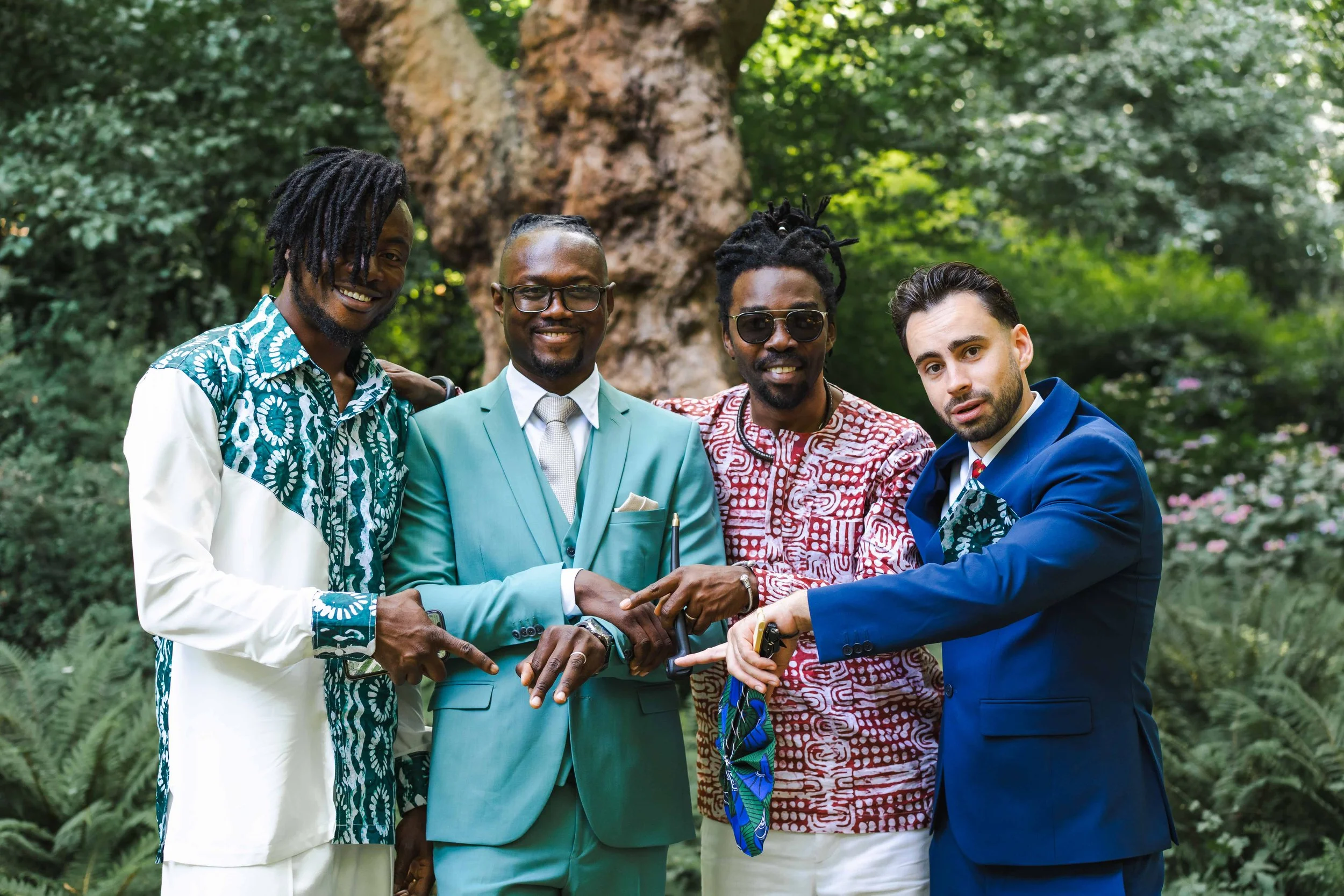 Cinq hommes habillés élégamment, posant ensemble dans un jardin avec un arbre en arrière-plan, manifestant de la camaraderie et de la confiance.