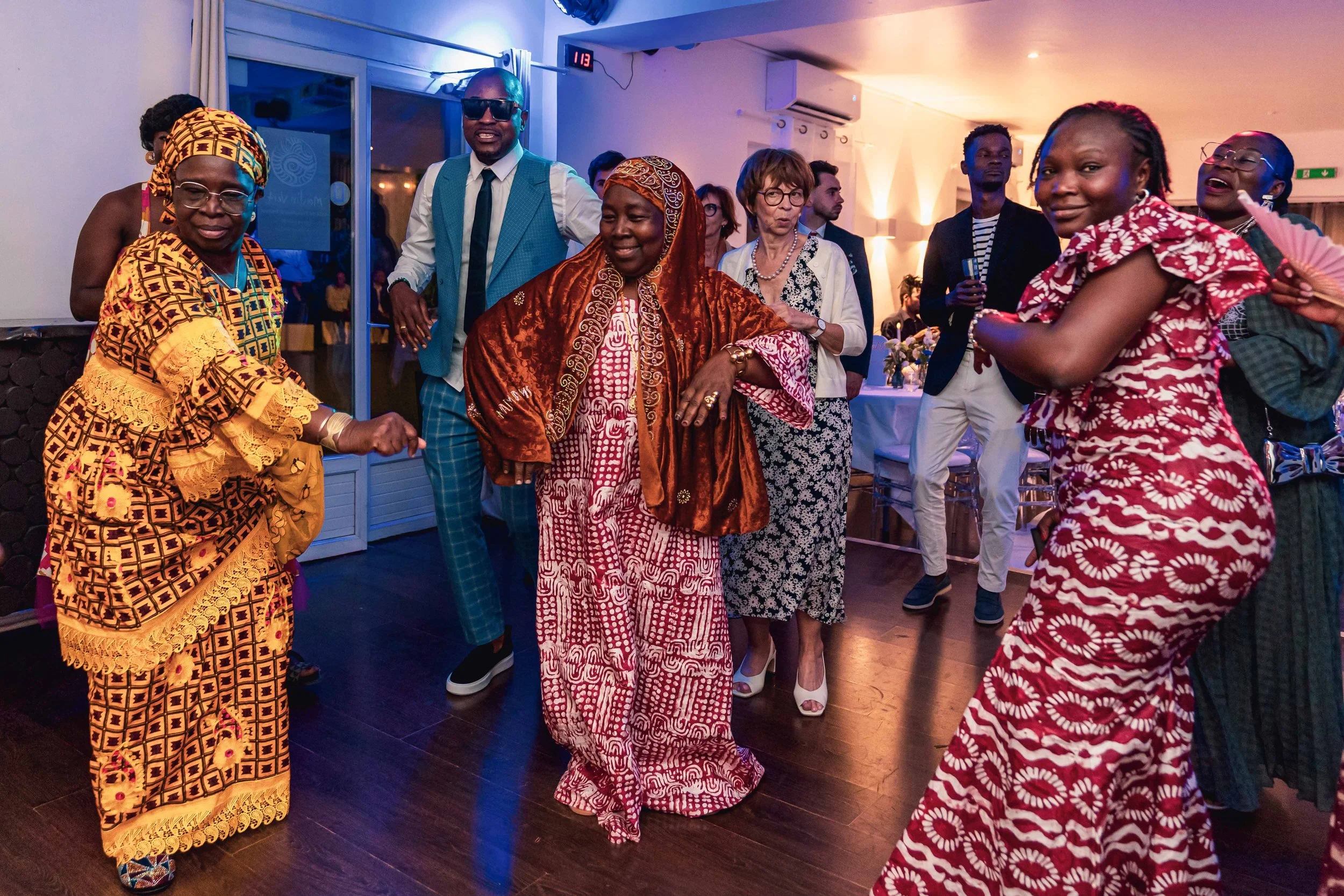 Groupe de personnes dansant et souriant à une fête, vêtues de tenues colorées et traditionnelles, ambiance festive avec éclairage chaud et décorations élégantes.