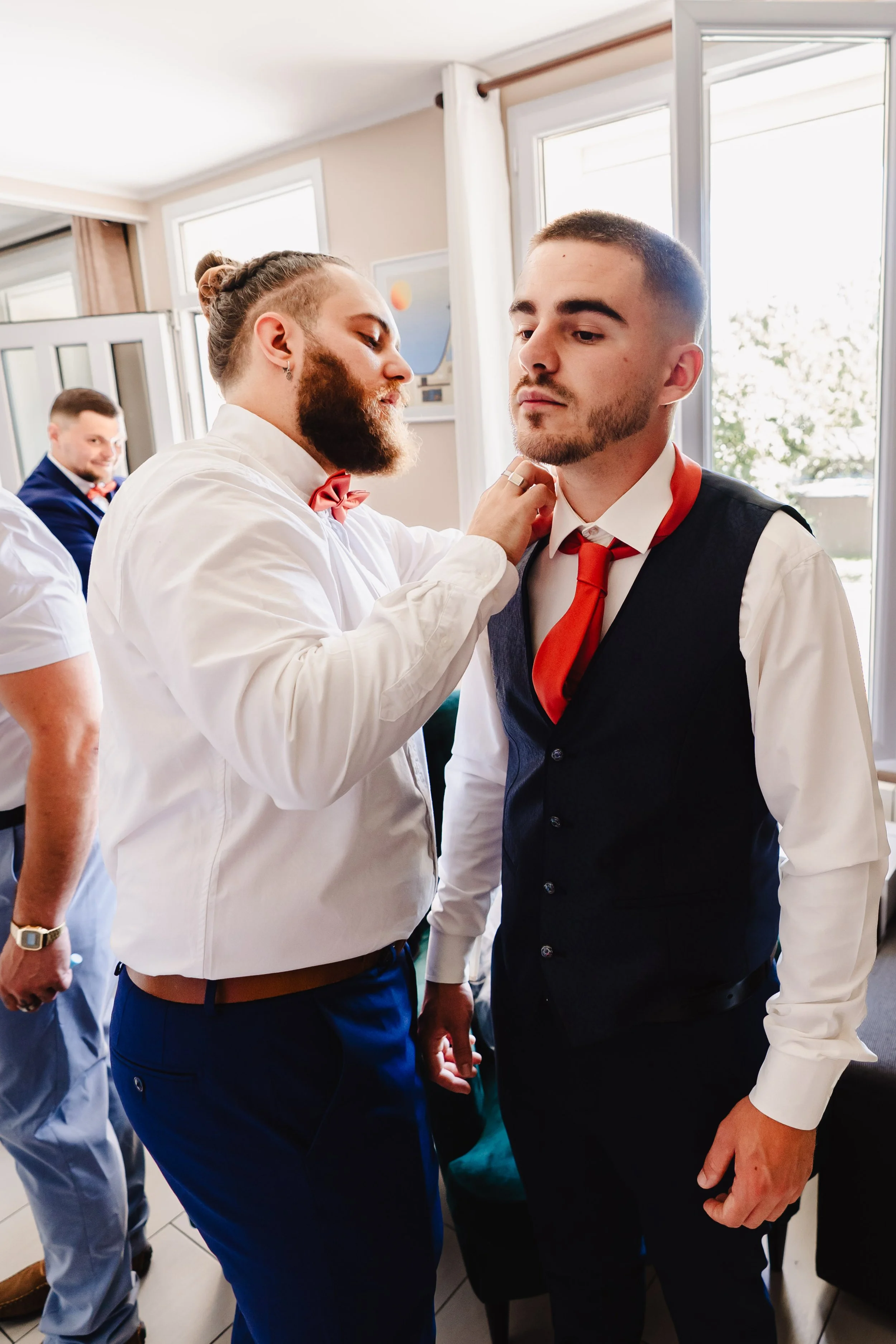 Deux hommes habillés en costume ajustent la cravate de l'autre dans une pièce lumineuse avec fenêtres, un autre homme en arrière-plan sourit.