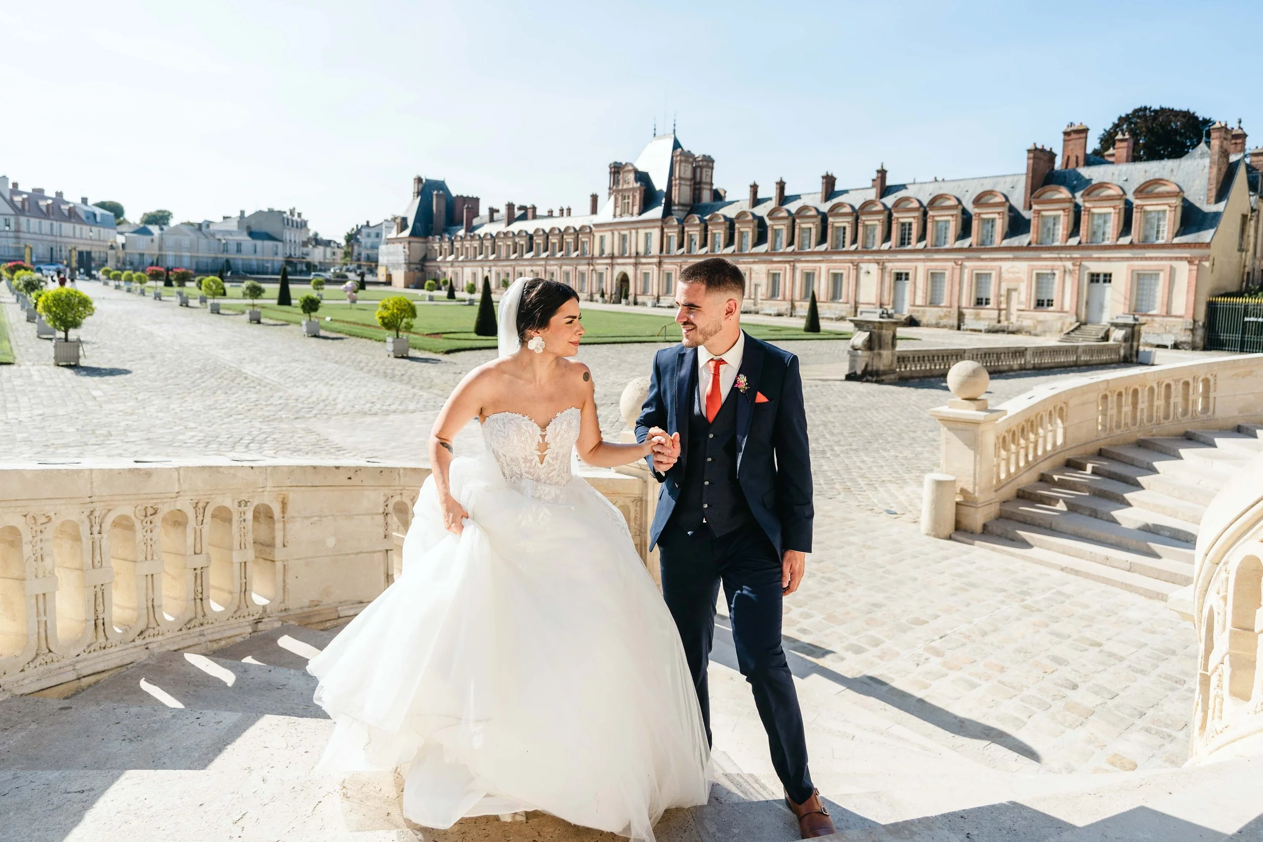 Un couple de mariés devant le château de Fontainebleau, en costume de mariage et en robe blanche, se tenant la main sur un escalier en pierre, par une journée ensoleillée.