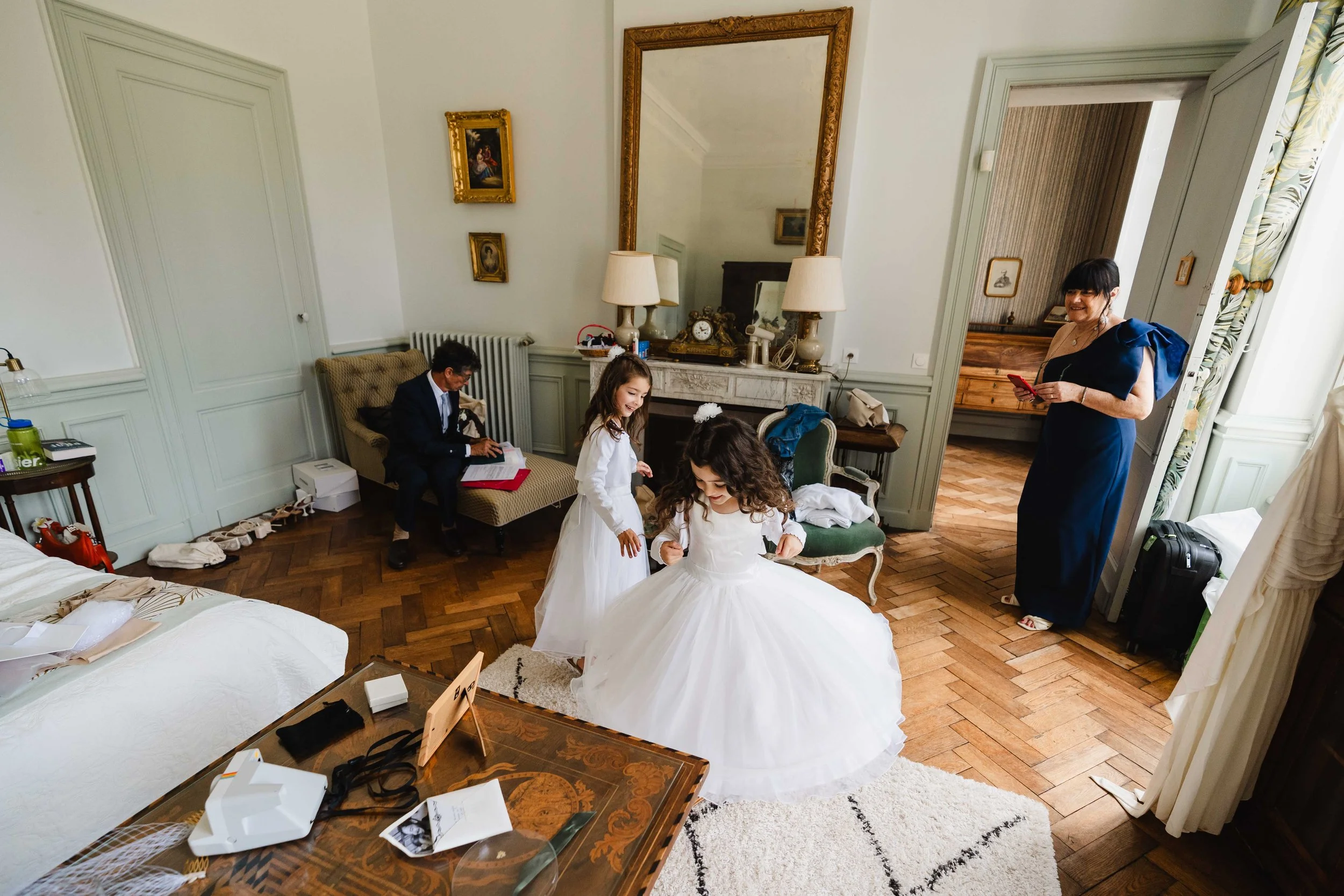 Une scène dans une chambre élégante où deux jeunes filles en robes de demoiselle d'honneur gravité, une femme en robe de soirée en train de sourire, et un homme en costume assis en arrière-plan.