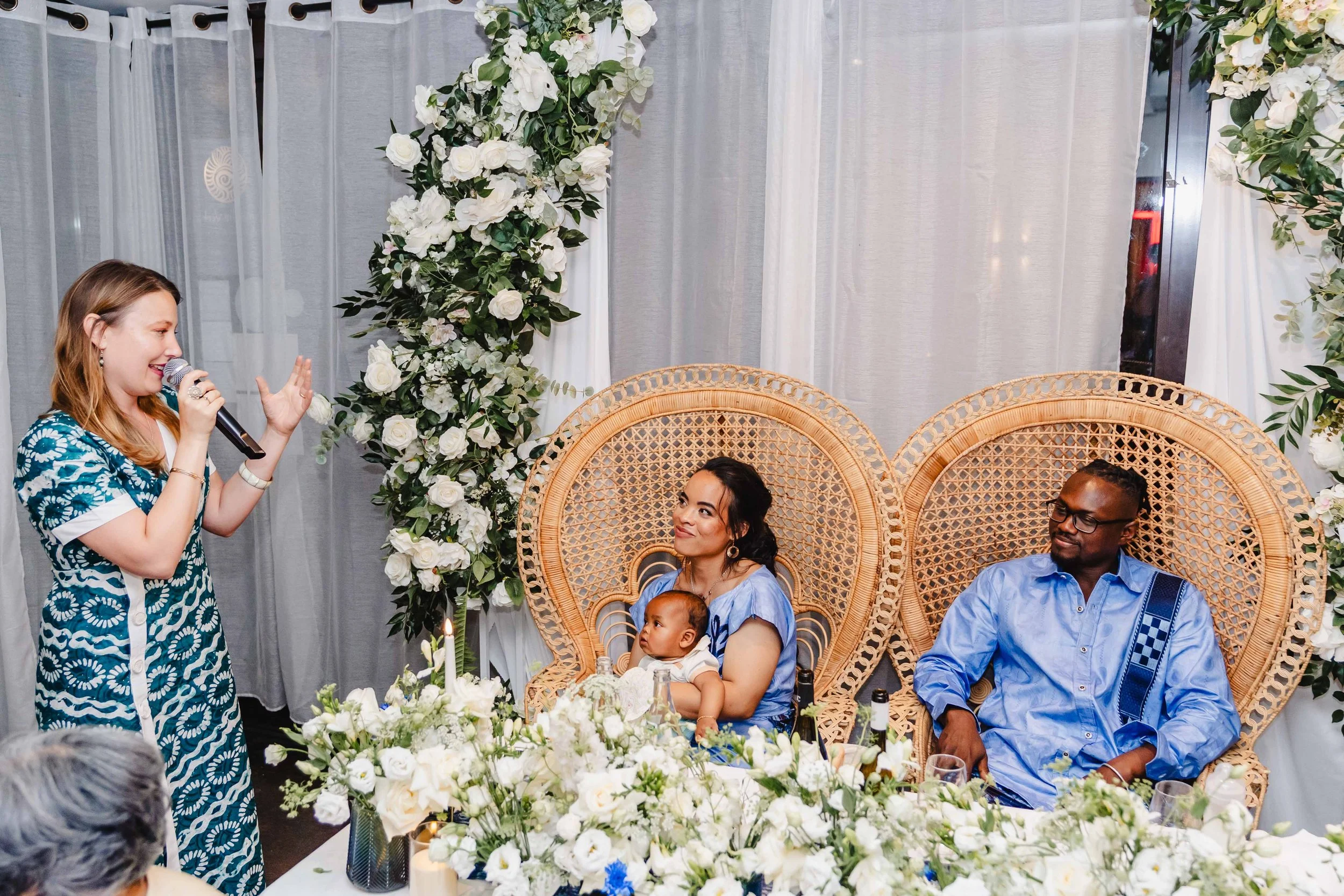 Une femme parle lors d'une cérémonie ou d'un repas, avec deux personnes assises sur des chaises en rotin tressé, décorées avec des fleurs blanches, une femme tenant un bébé et un homme portant des lunettes et une chemise bleue.