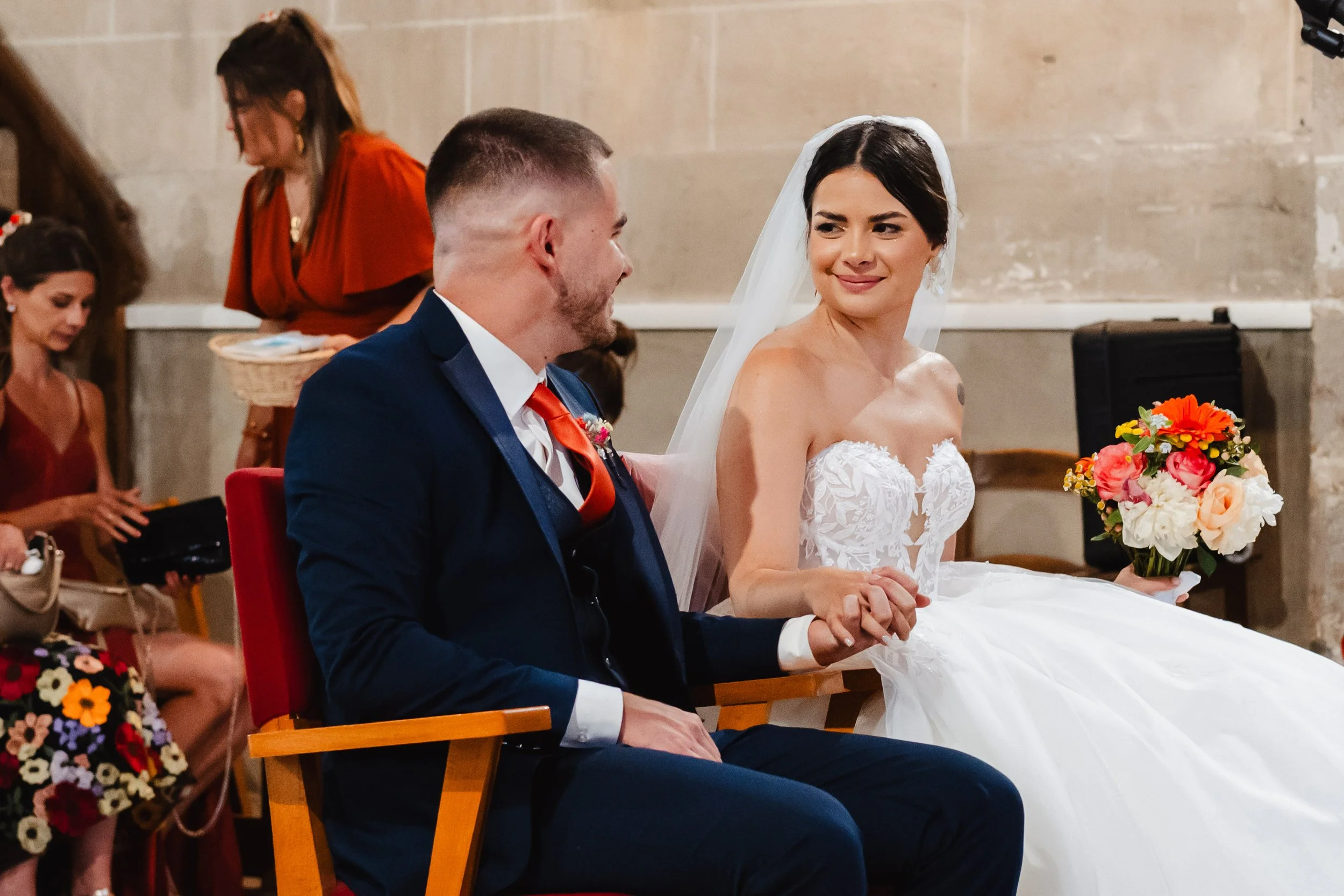 Un couple de mariés lors d'une cérémonie de mariage. La mariée porte une robe blanche avec un voile et tient un bouquet de fleurs colorées. L'homme porte un costume bleu foncé avec une cravate orange. Ils se tiennent la main et se regardent avec tend