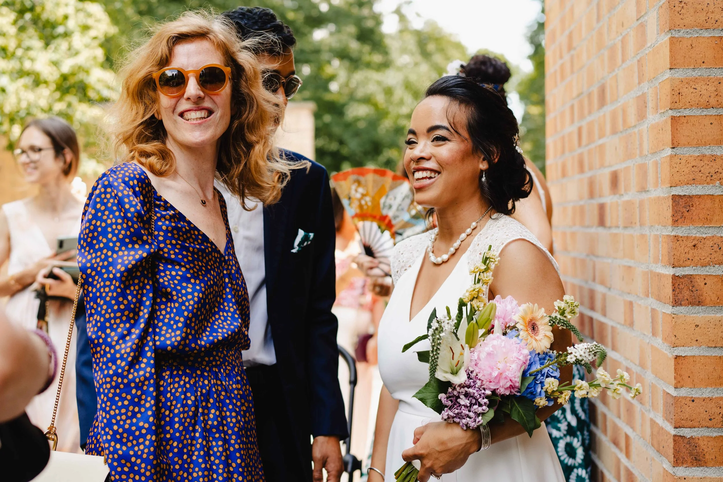 Photo de mariage montrant trois femmes souriantes, avec une de la mariée qui tient un bouquet de fleurs, en train de discuter à l'extérieur, près d'un mur en briques.