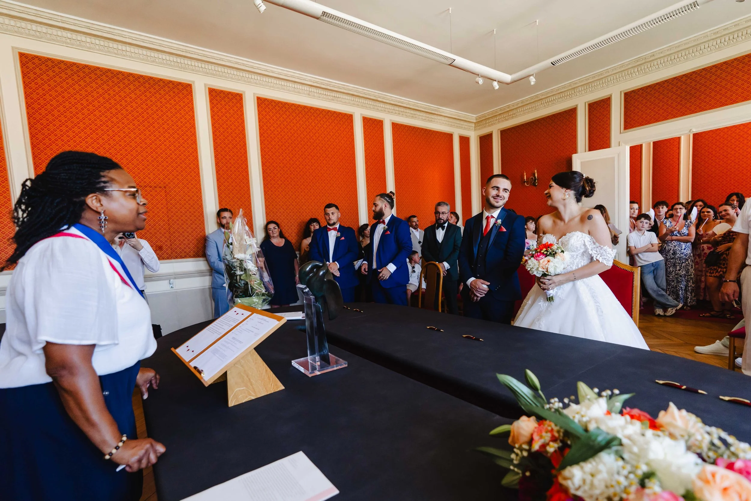 Un couple de mariés lors de leur cérémonie de mariage, entourés de témoins et de proches dans la salle de mariage de Bagneux Sur Loing ornée de murs orange et de moulures blanches.