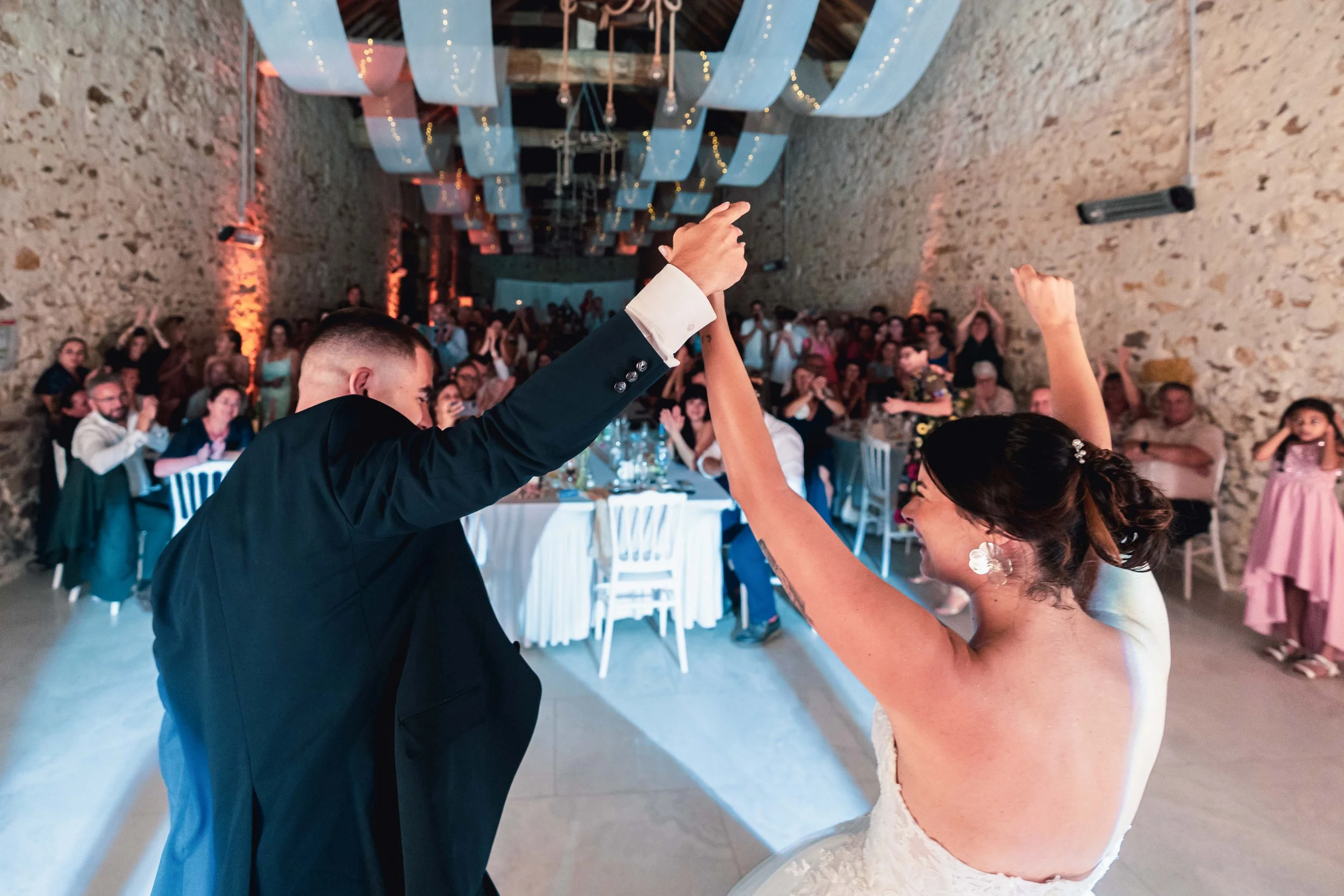 Un couple en costume de mariage danse lors d'une cérémonie dans la salle de réception du domaine de Mauperthuis décorée pour un mariage, avec un public assis et applaudissant en arrière-plan.
